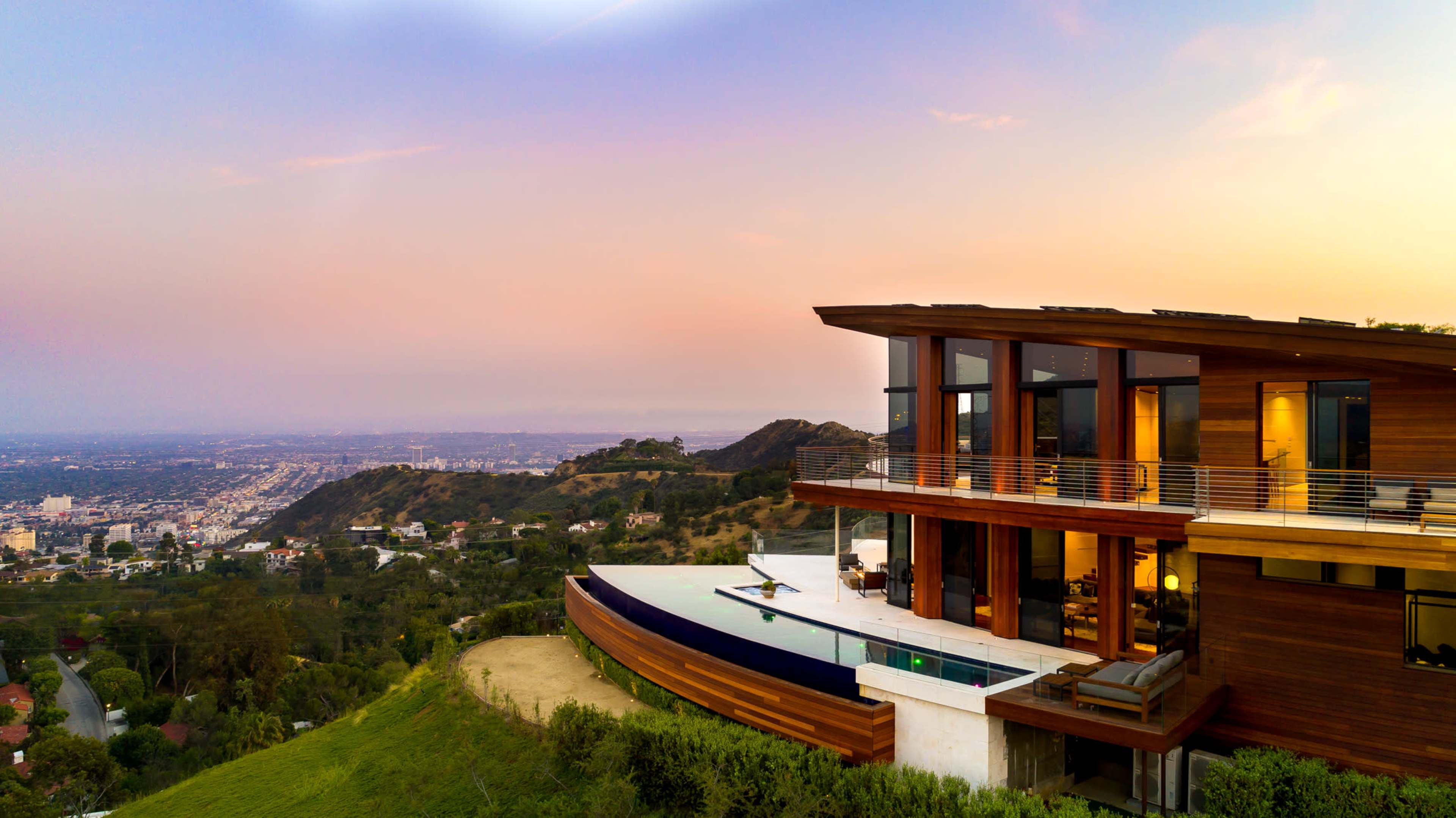 A modern house with large windows overlooks a city skyline at sunset, set against a hillside.
