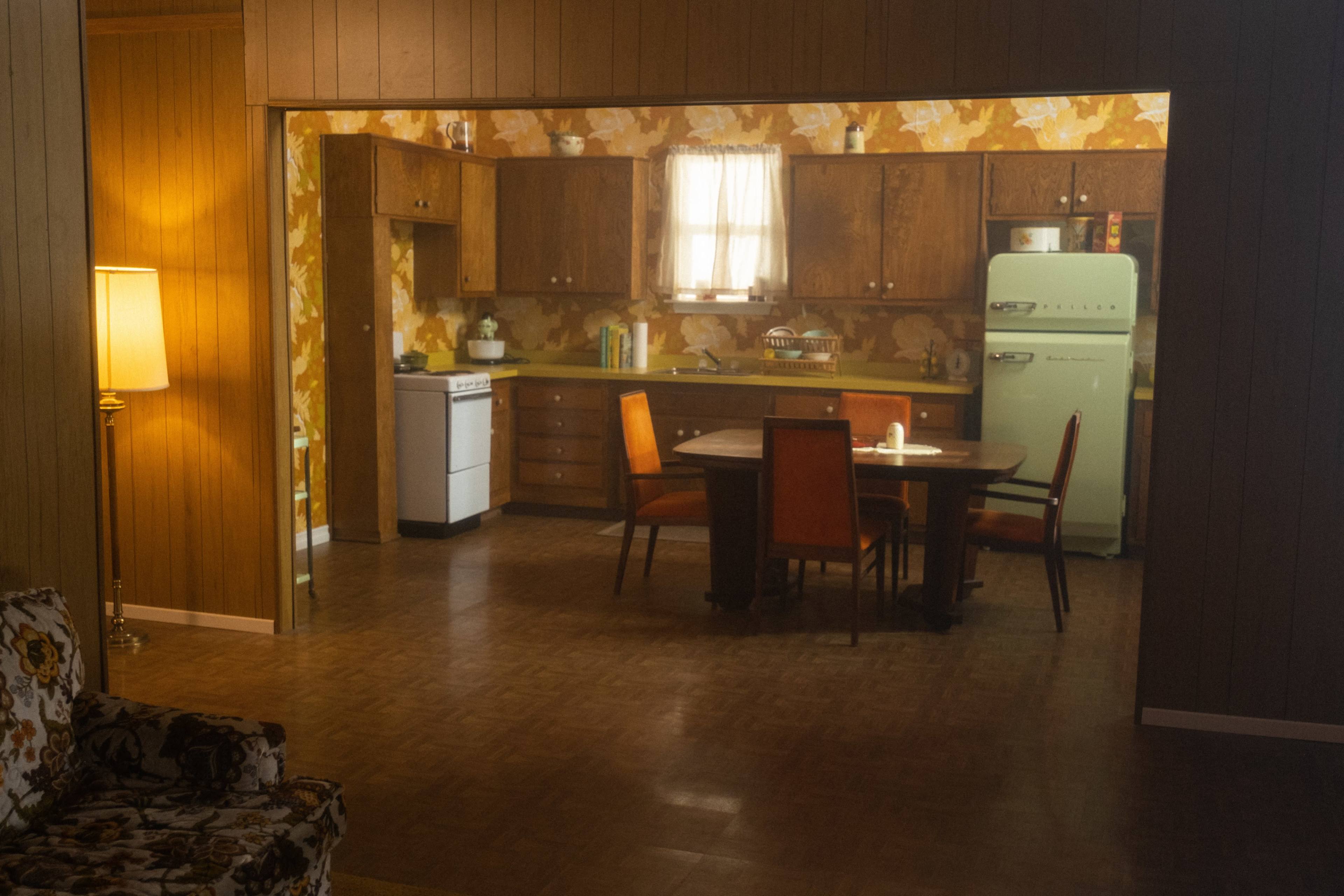 The image shows a retro kitchen with wooden cabinets, an old-fashioned stove, and a green refrigerator, as viewed from a living room with patterned furniture.