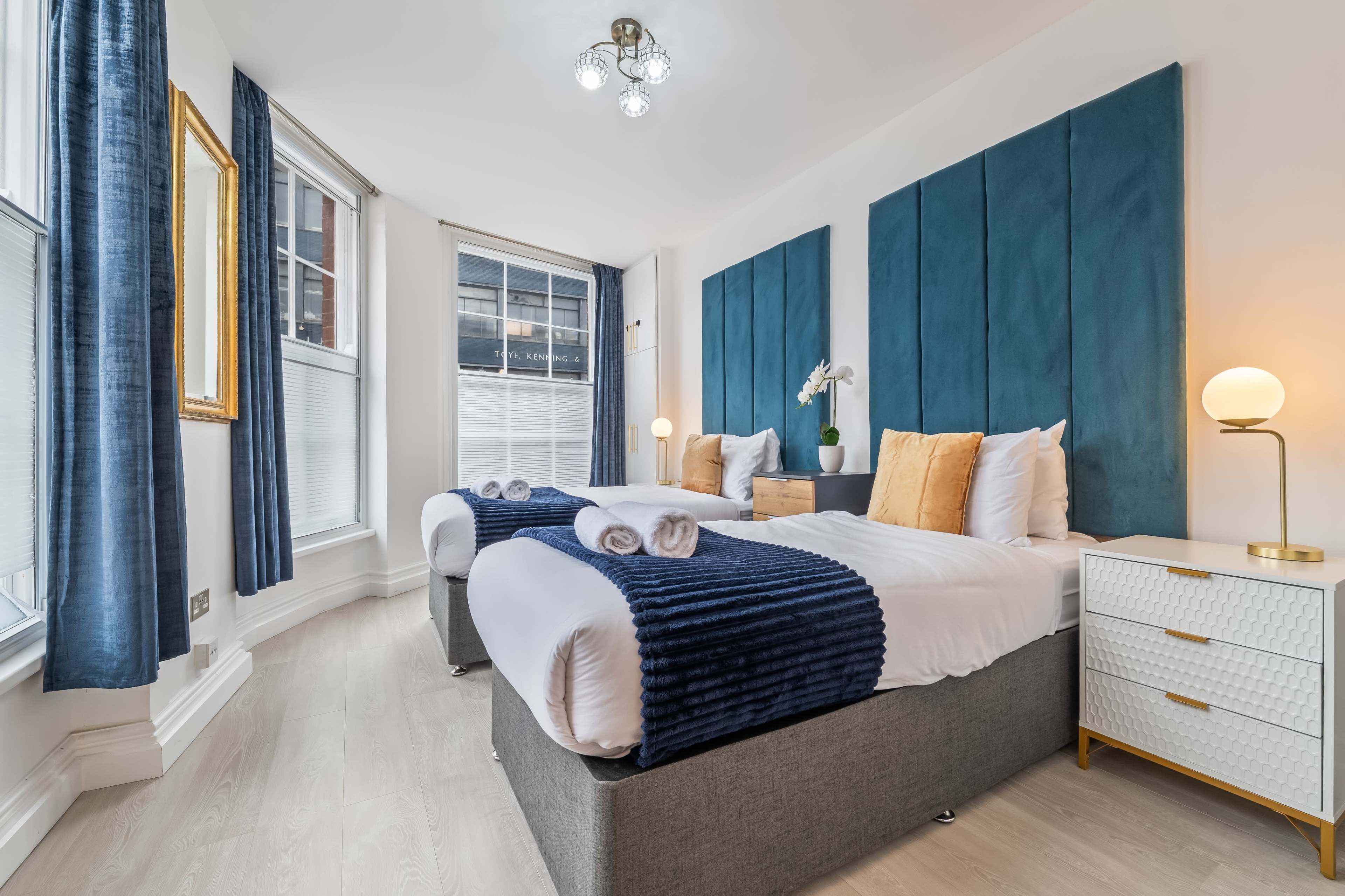 The image shows a neatly arranged hotel room with two beds, blue accent walls, and modern furniture.