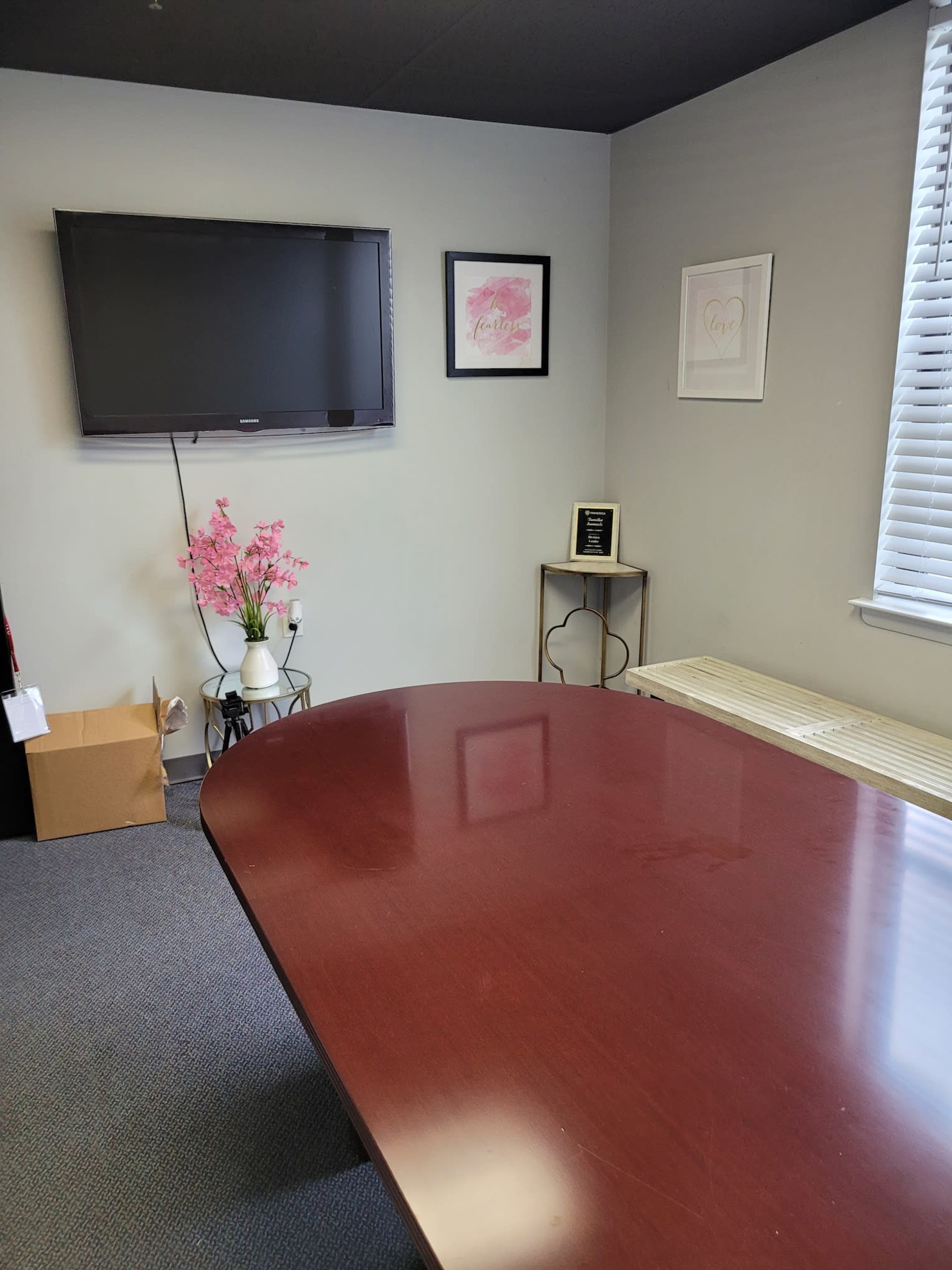 A conference room with a large wooden table, a wall-mounted television, framed artwork, and a decorative plant near the corner.