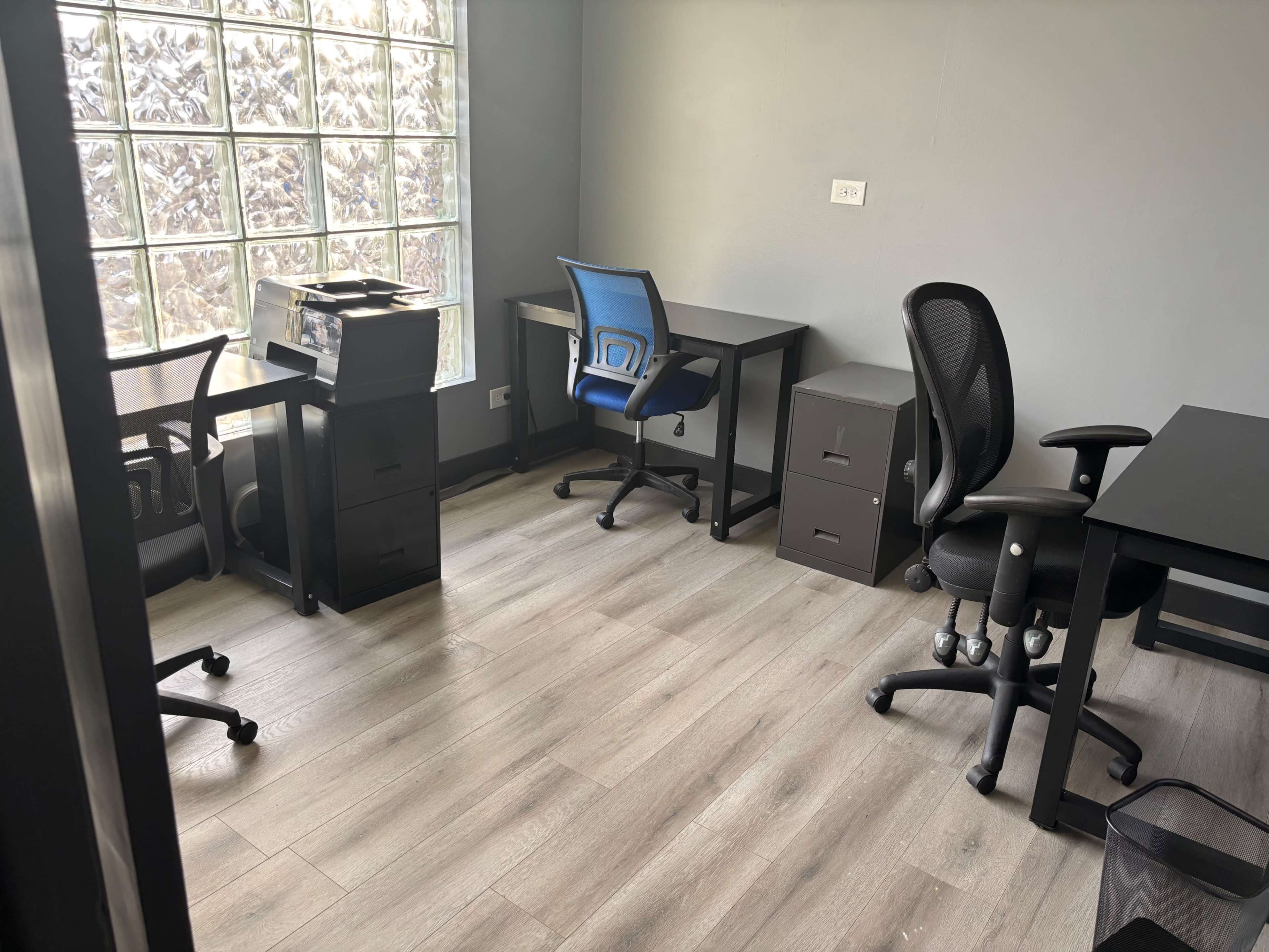 The image shows a modern office space featuring three desks, two office chairs, a printer, and a filing cabinet, all arranged on a wooden floor.