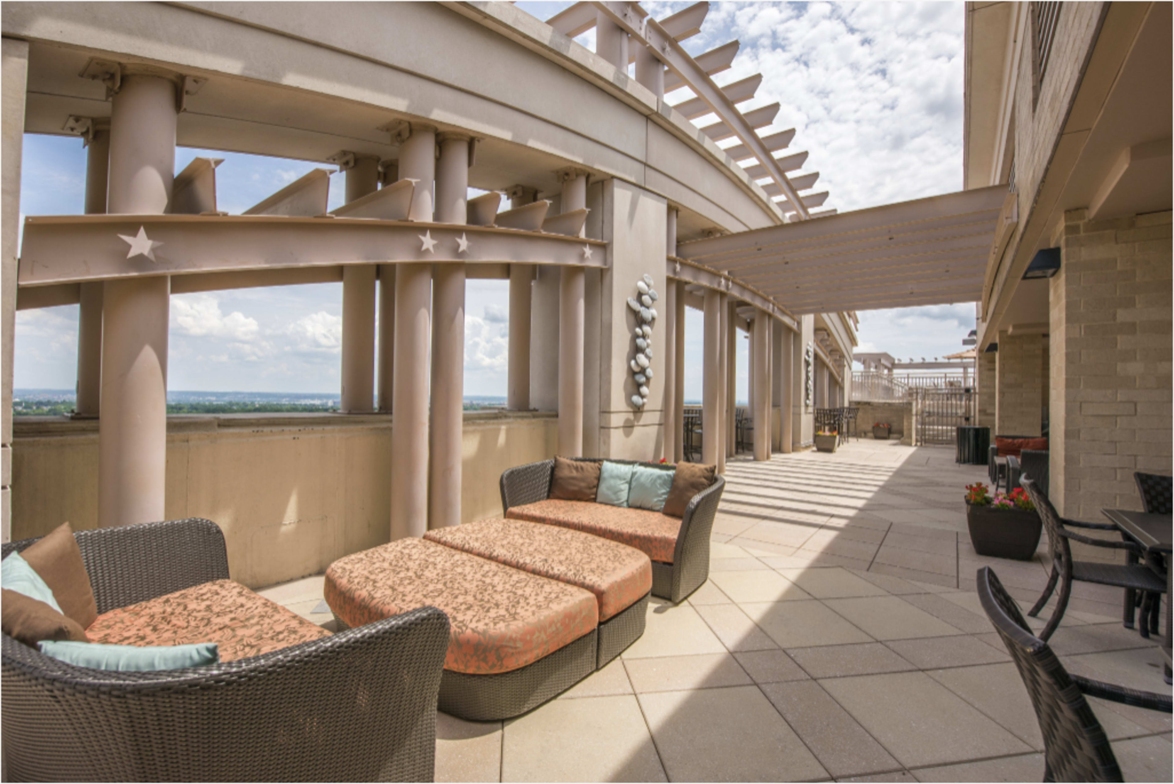 The image shows a spacious rooftop terrace with modern wicker seating and a view of the sky.