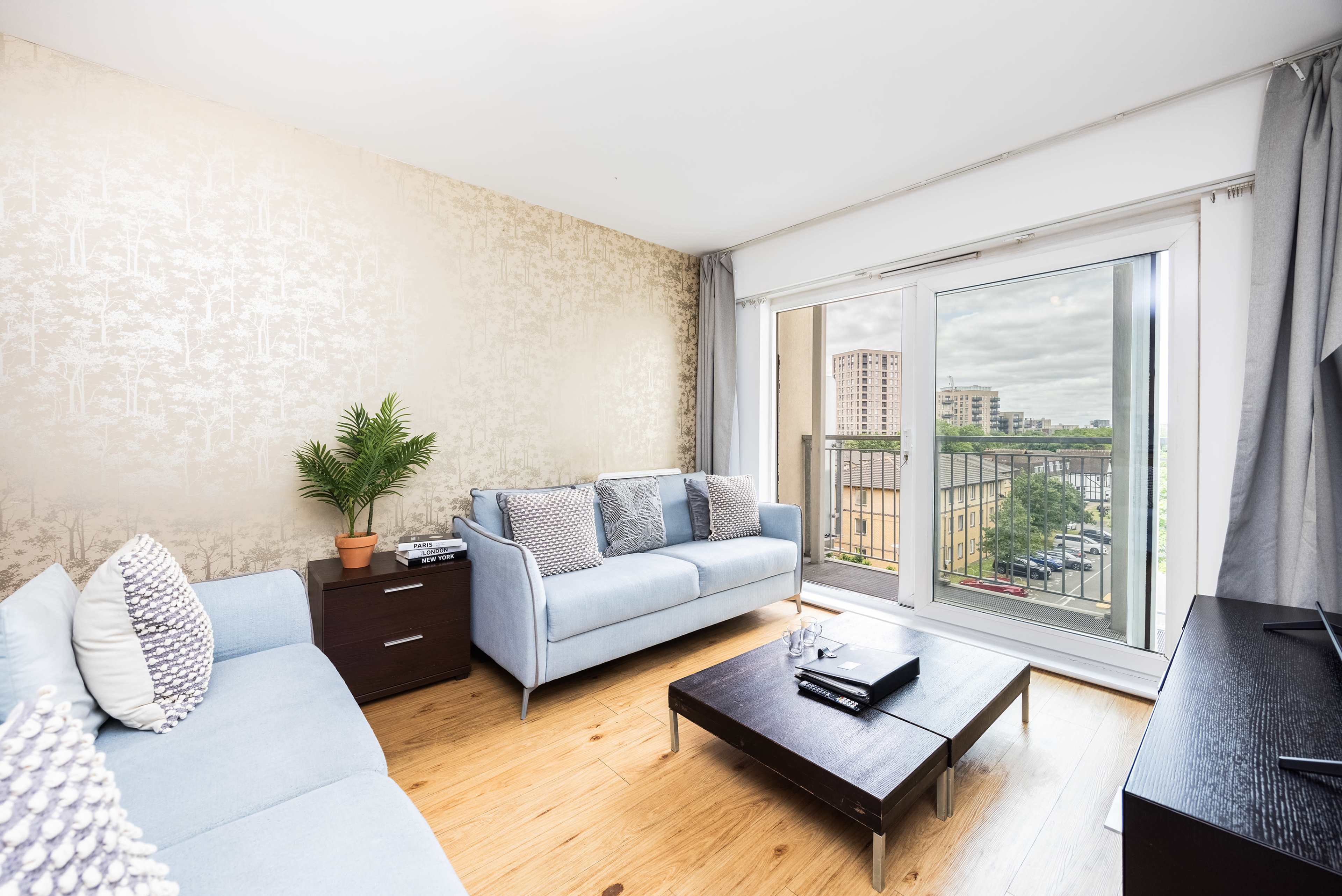 The living room features two light blue sofas, a dark wood coffee table, and a sliding glass door leading to a balcony with a view of buildings outside.