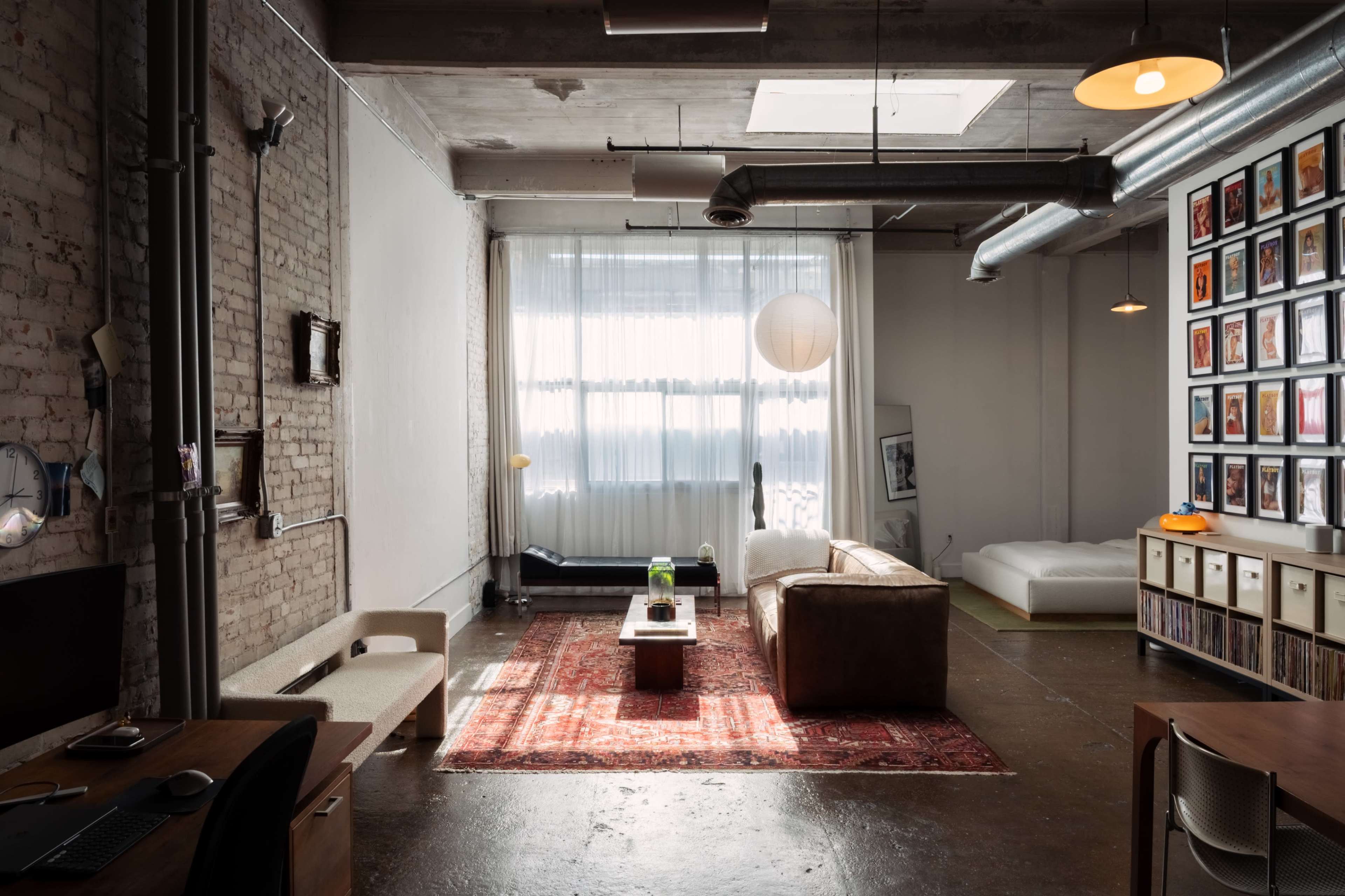 The image shows a spacious loft-style living area with exposed brick walls, large windows, a brown sofa, and a gallery wall of framed photos.