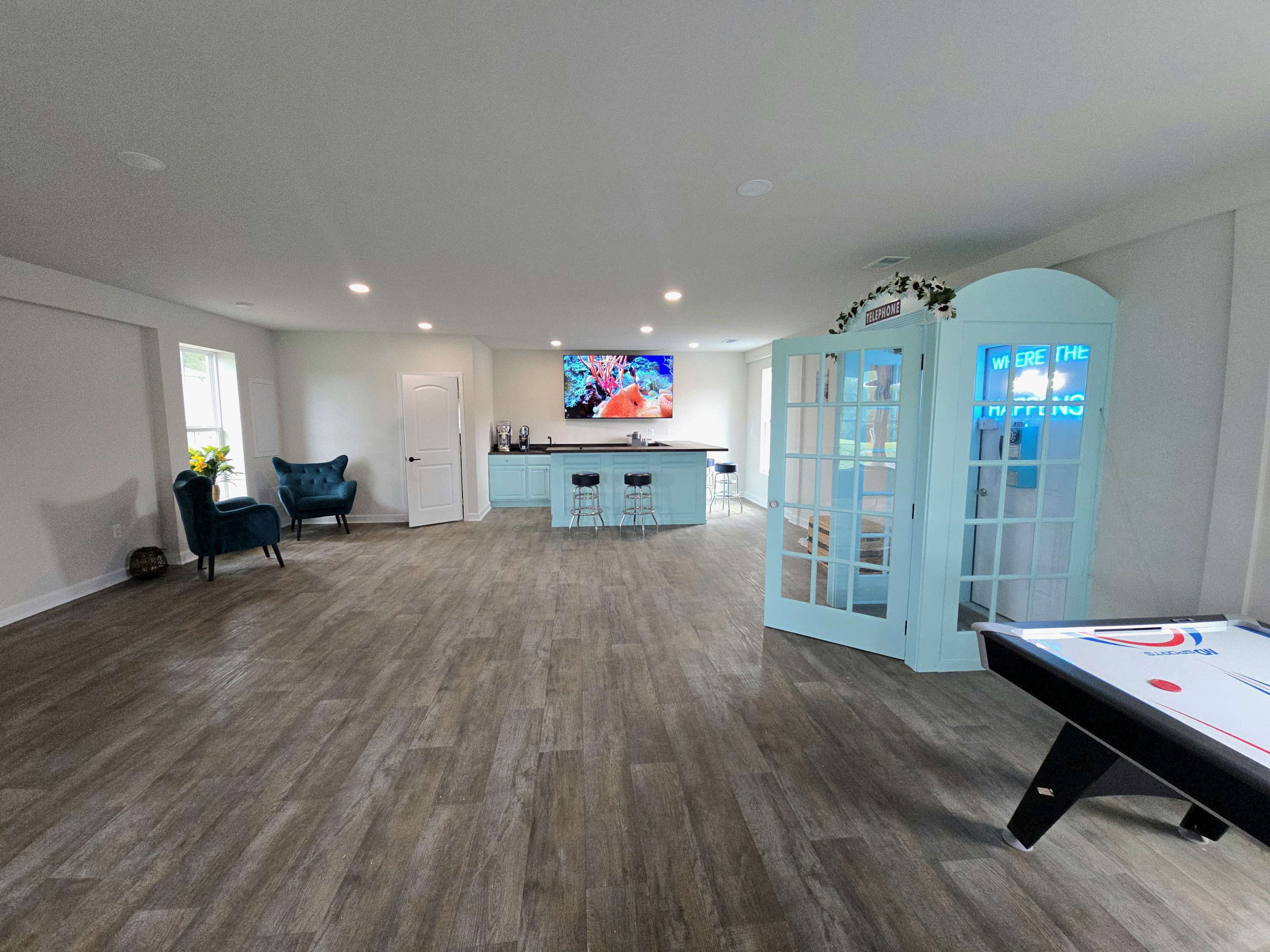 The image shows a spacious, modern game room with a bar area, a large wall-mounted TV, a pool table, and a blue telephone booth-style structure.