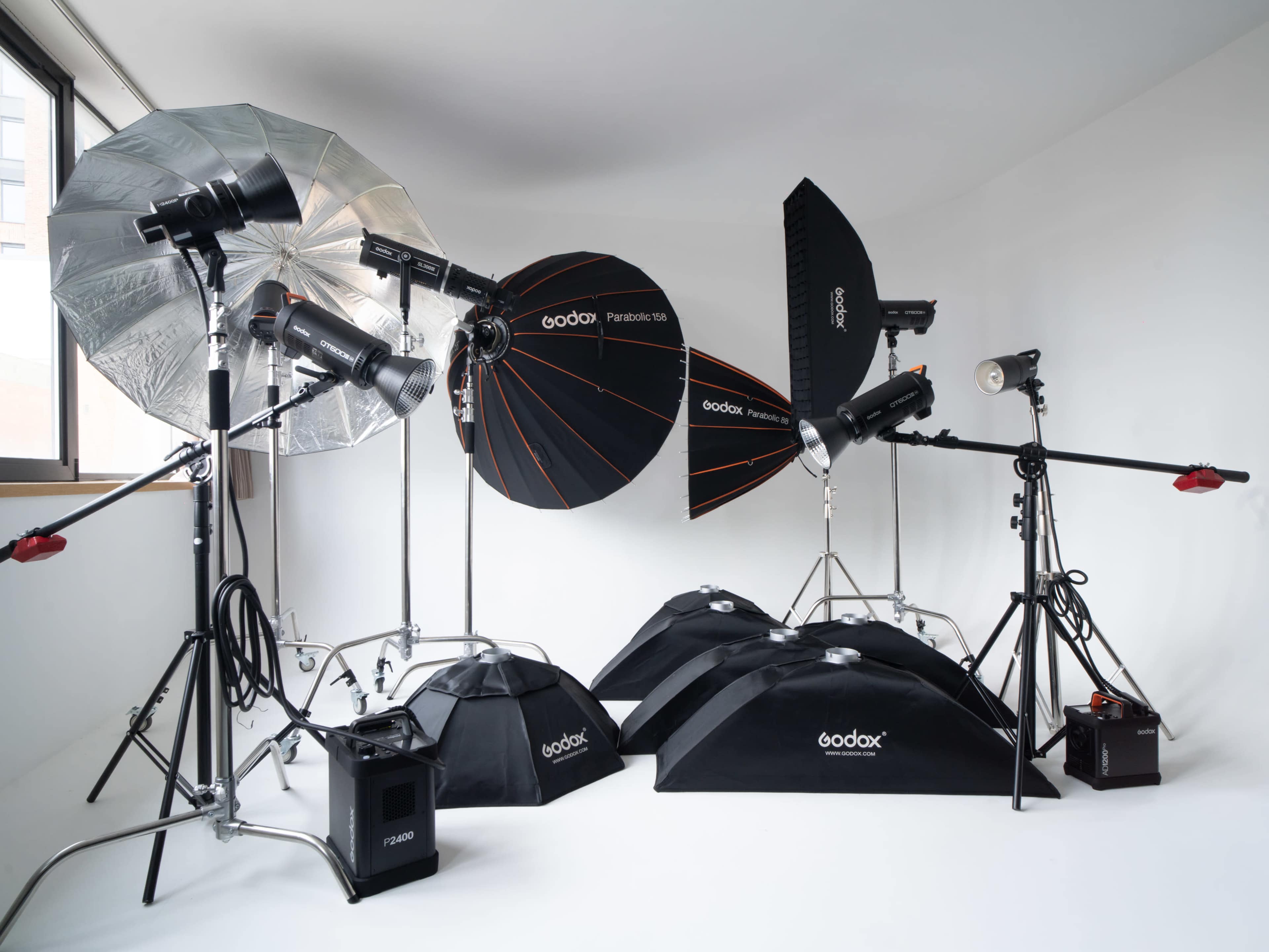 The image shows a photography studio setup featuring multiple light sources, softboxes, and light modifiers arranged on stands against a white backdrop.