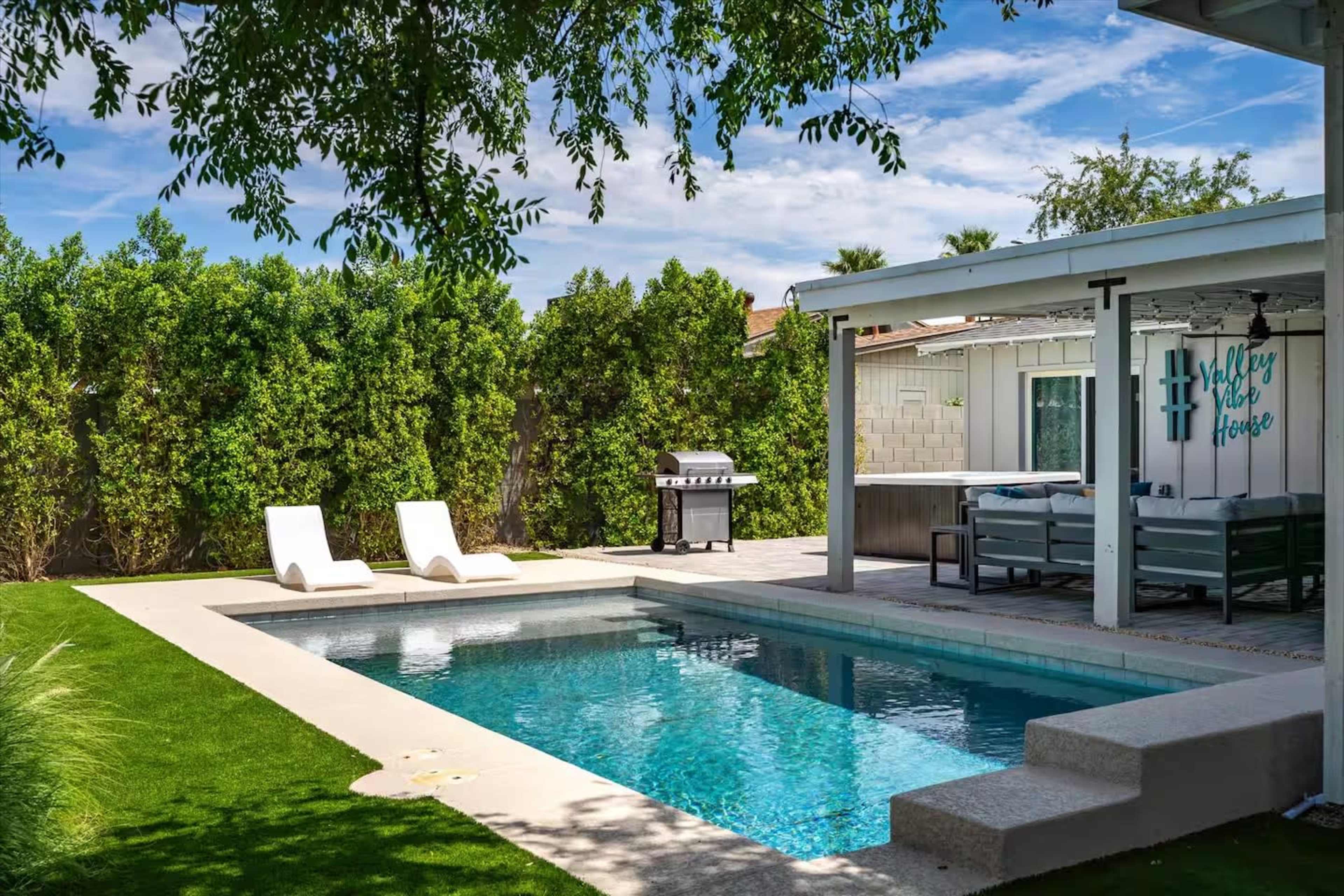 The image shows a backyard with a swimming pool, two lounge chairs, and a covered patio area with a barbecue grill and seating.