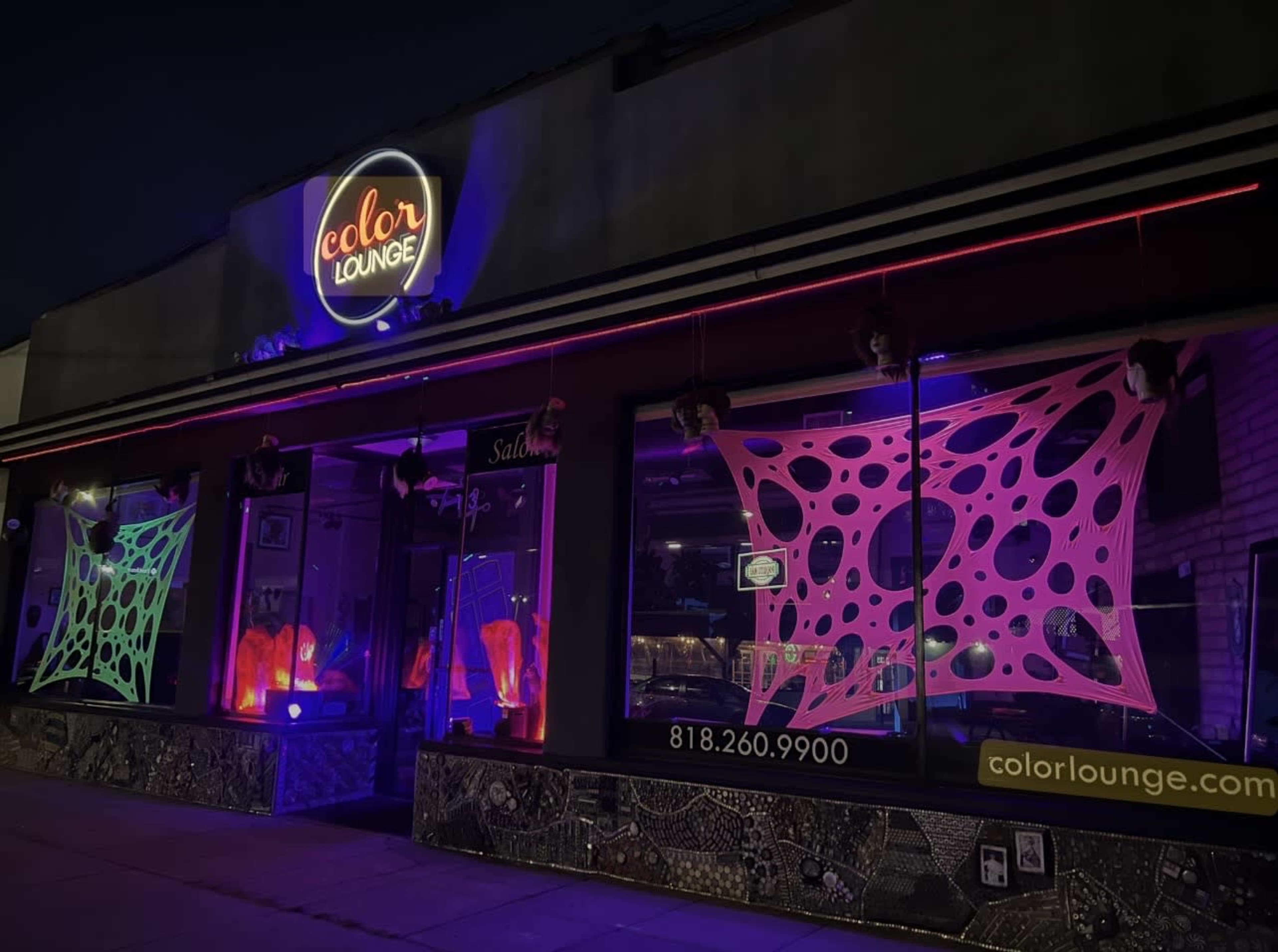 The storefront of Color Lounge features bright neon lights and colorful decorations on its windows, with a clear night sky in the background.