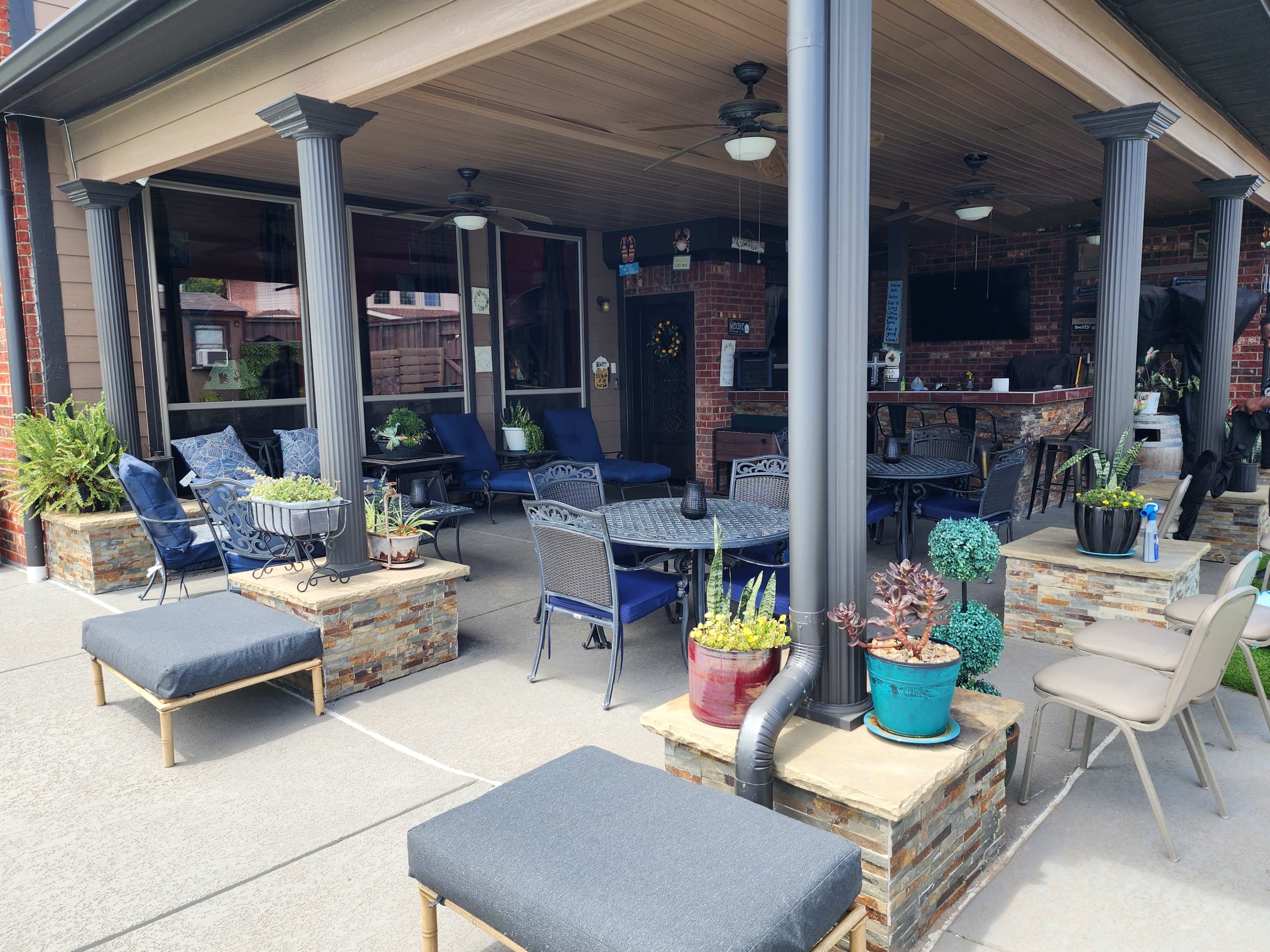 The image features a covered patio area with seating, stone planters, and a television mounted on the wall, surrounded by decorative plants.