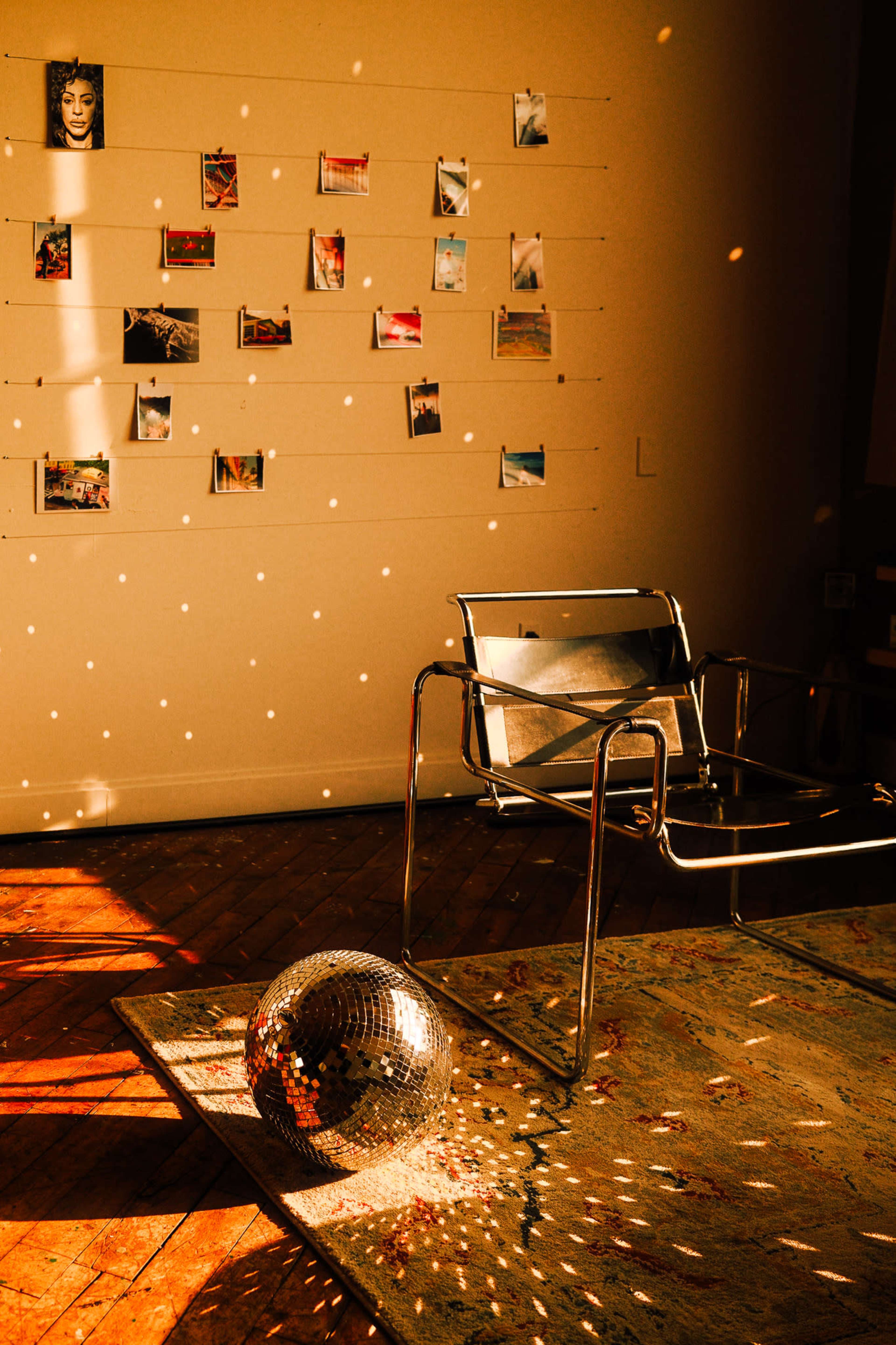 A modern chair sits on a rug beside a disco ball on the floor, with photographs displayed on a wall illuminated by warm sunlight.
