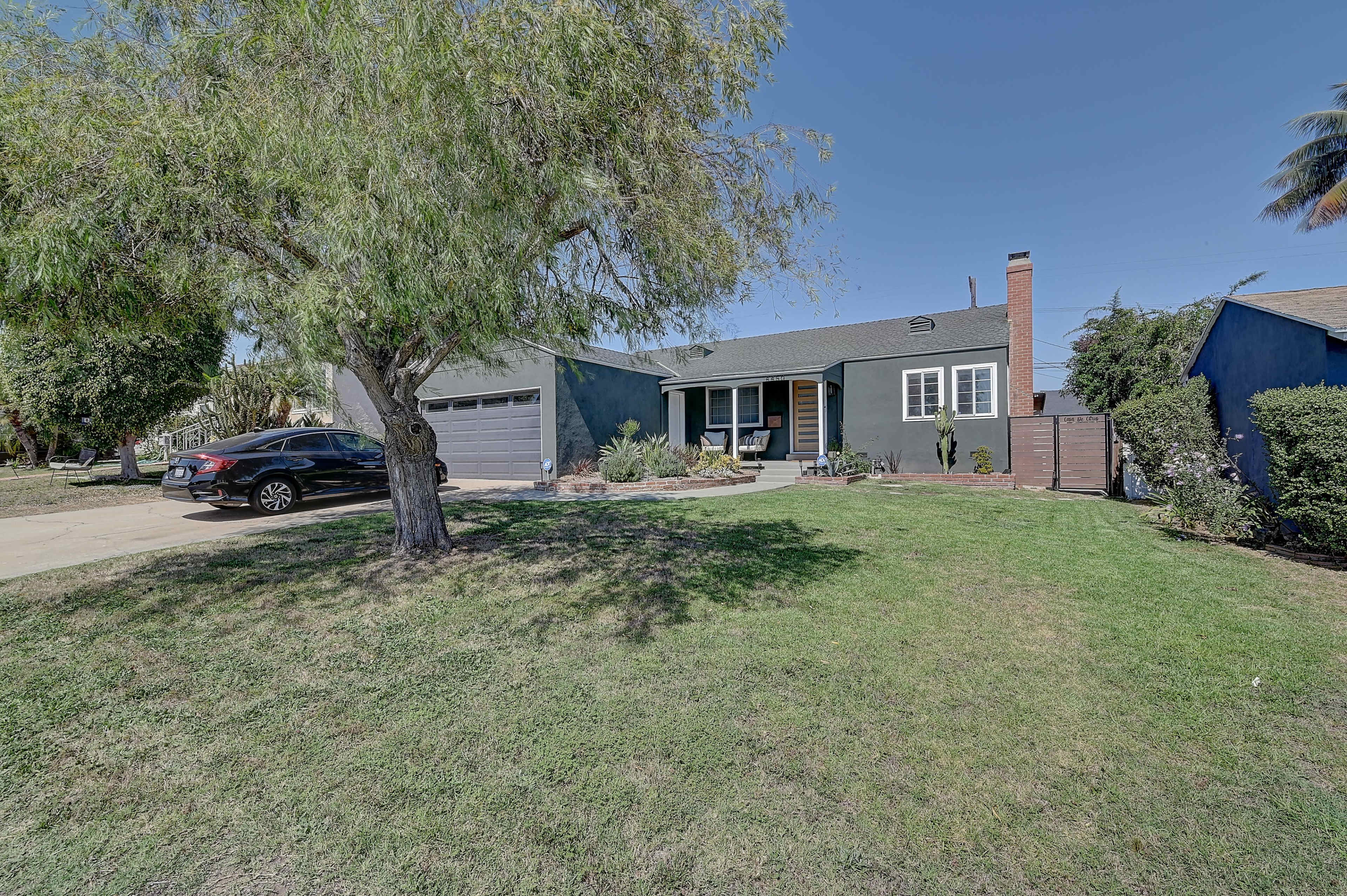 A single-story house with dark exterior paint, a driveway on the left, and a large front lawn featuring a tree.