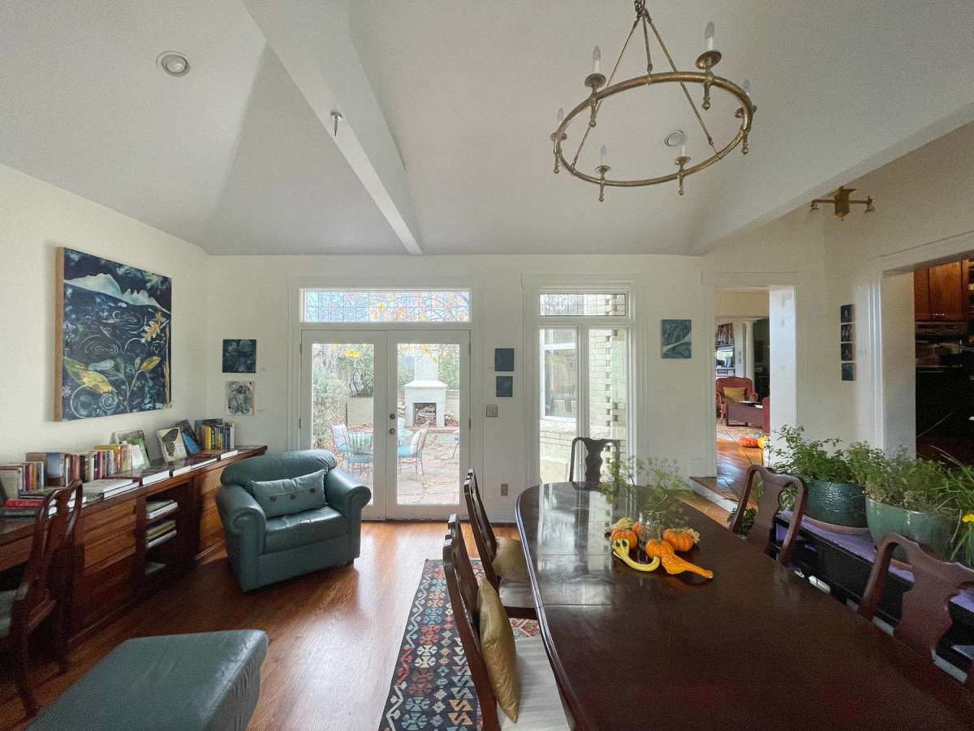 A dining room features a long wooden table surrounded by chairs, with a blue armchair and large windows that provide a view of an outdoor patio.
