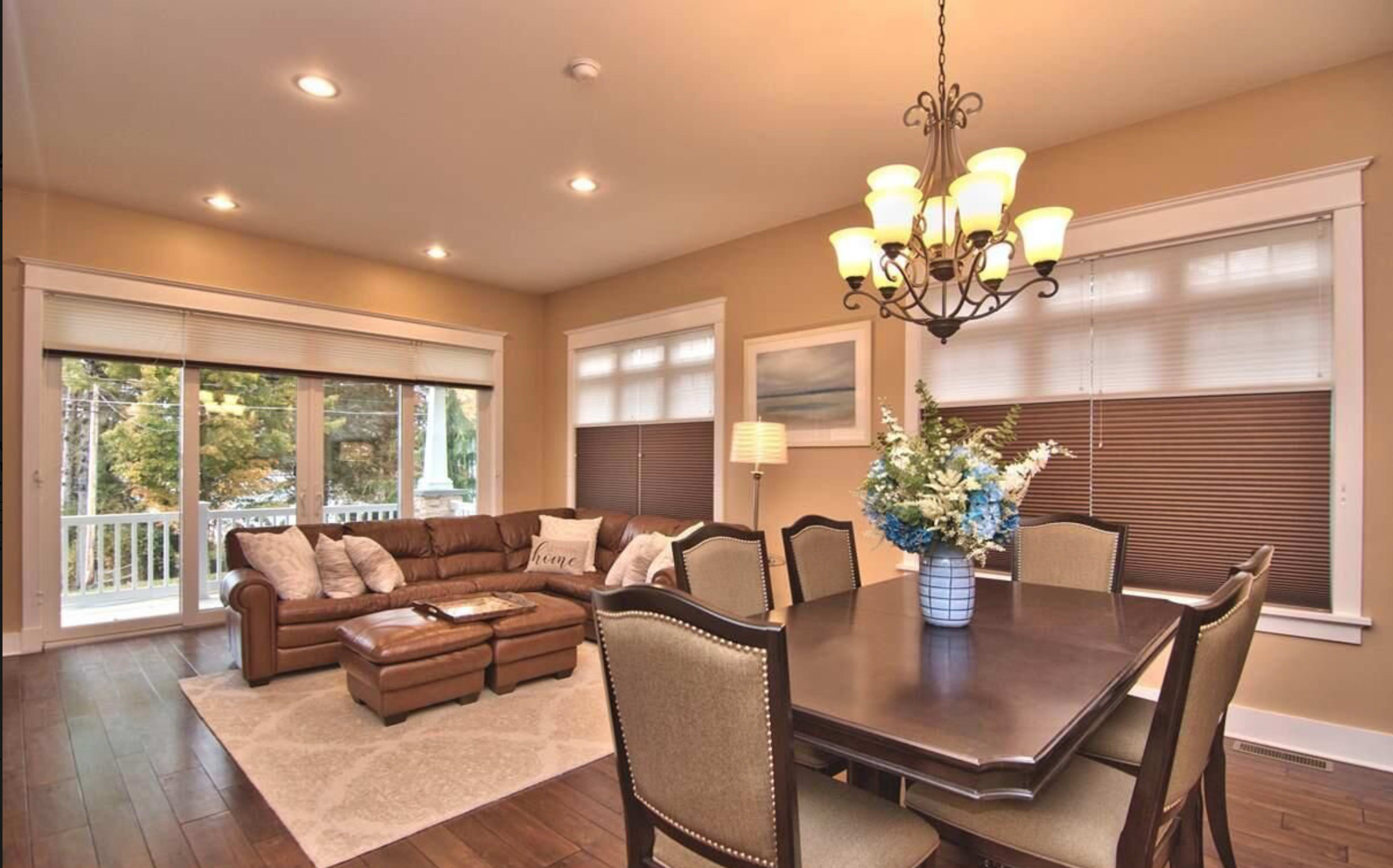 A cozy living and dining area featuring a brown leather sofa, a wooden dining table with chairs, and large windows letting in natural light.