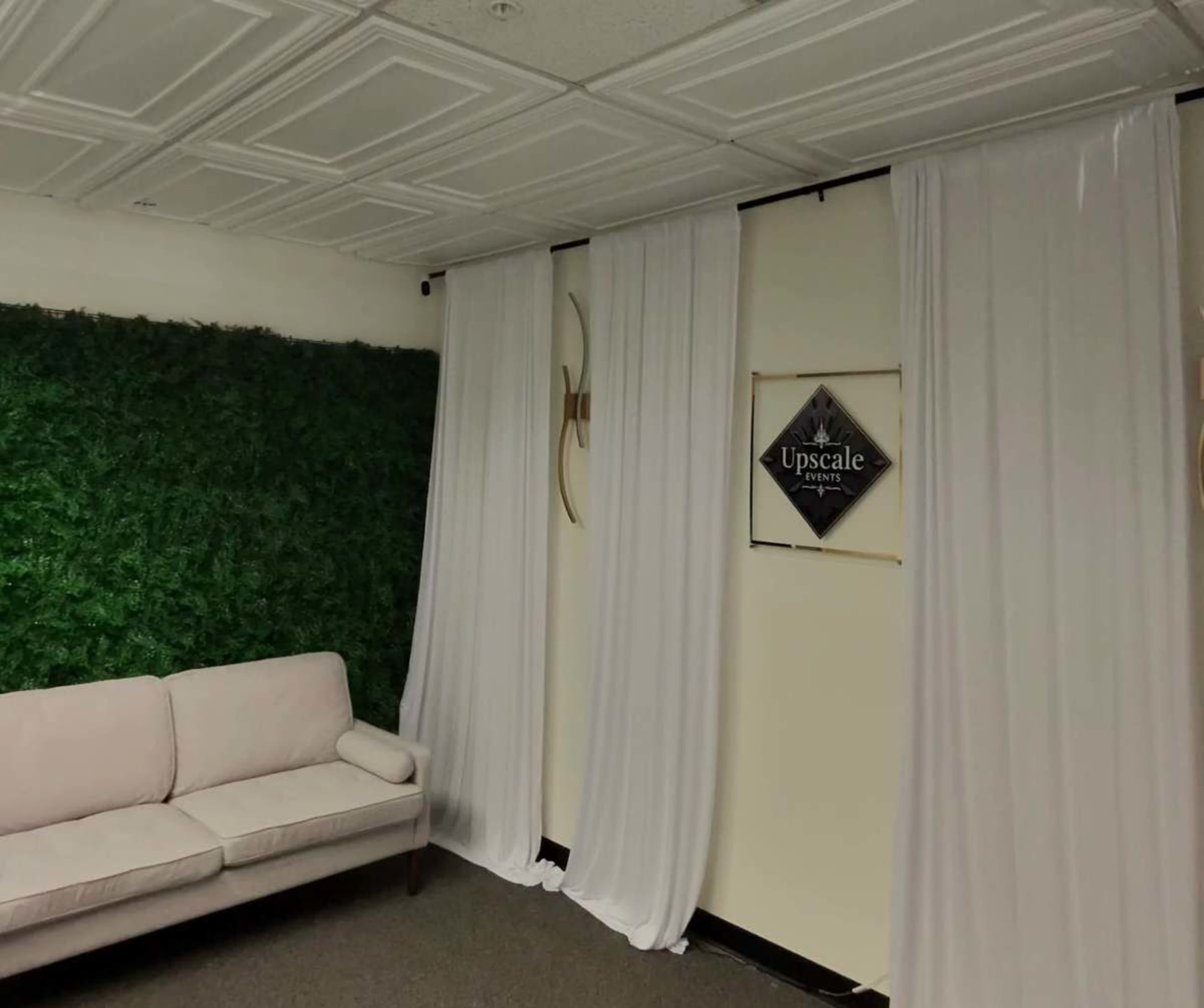 The image shows a room with a green wall made of artificial foliage, a white sofa, white drapery partitions, and a sign that reads "Upscale."