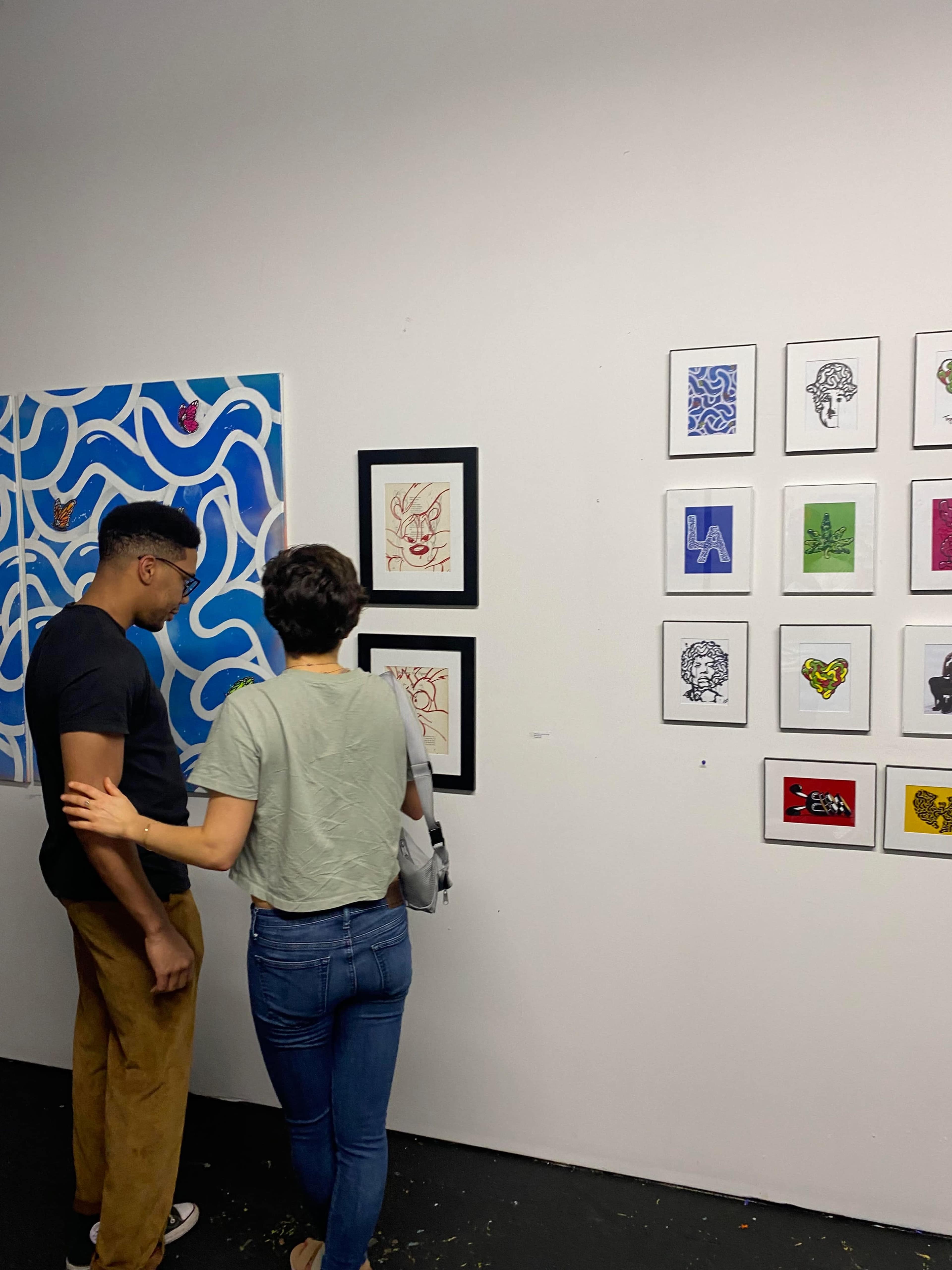 Two individuals stand in an art gallery, examining framed artworks displayed on the walls.
