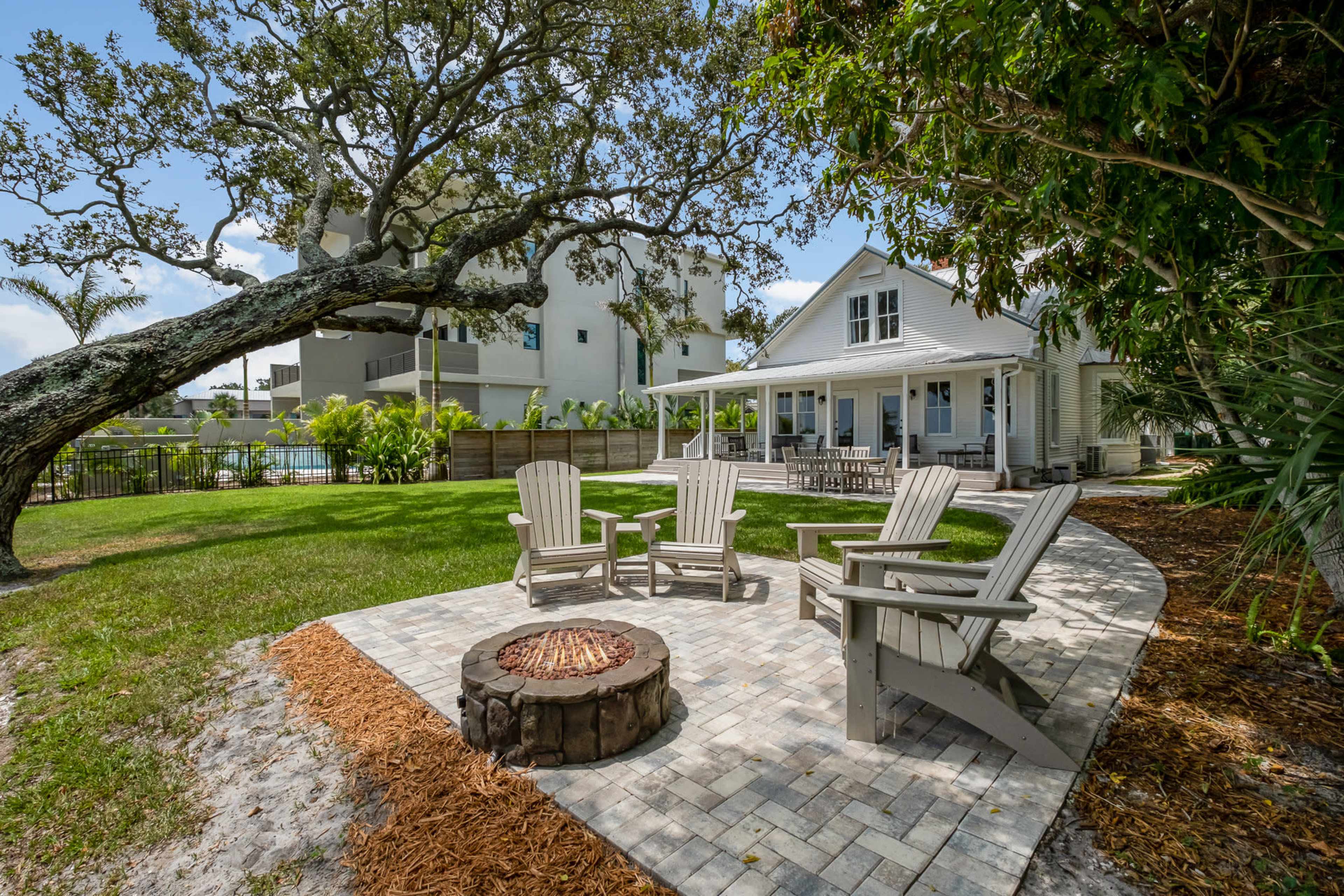 A backyard features a circular stone patio with four Adirondack chairs arranged around a fire pit, surrounded by lush greenery and a large tree, with a house and a high-rise building in the background.