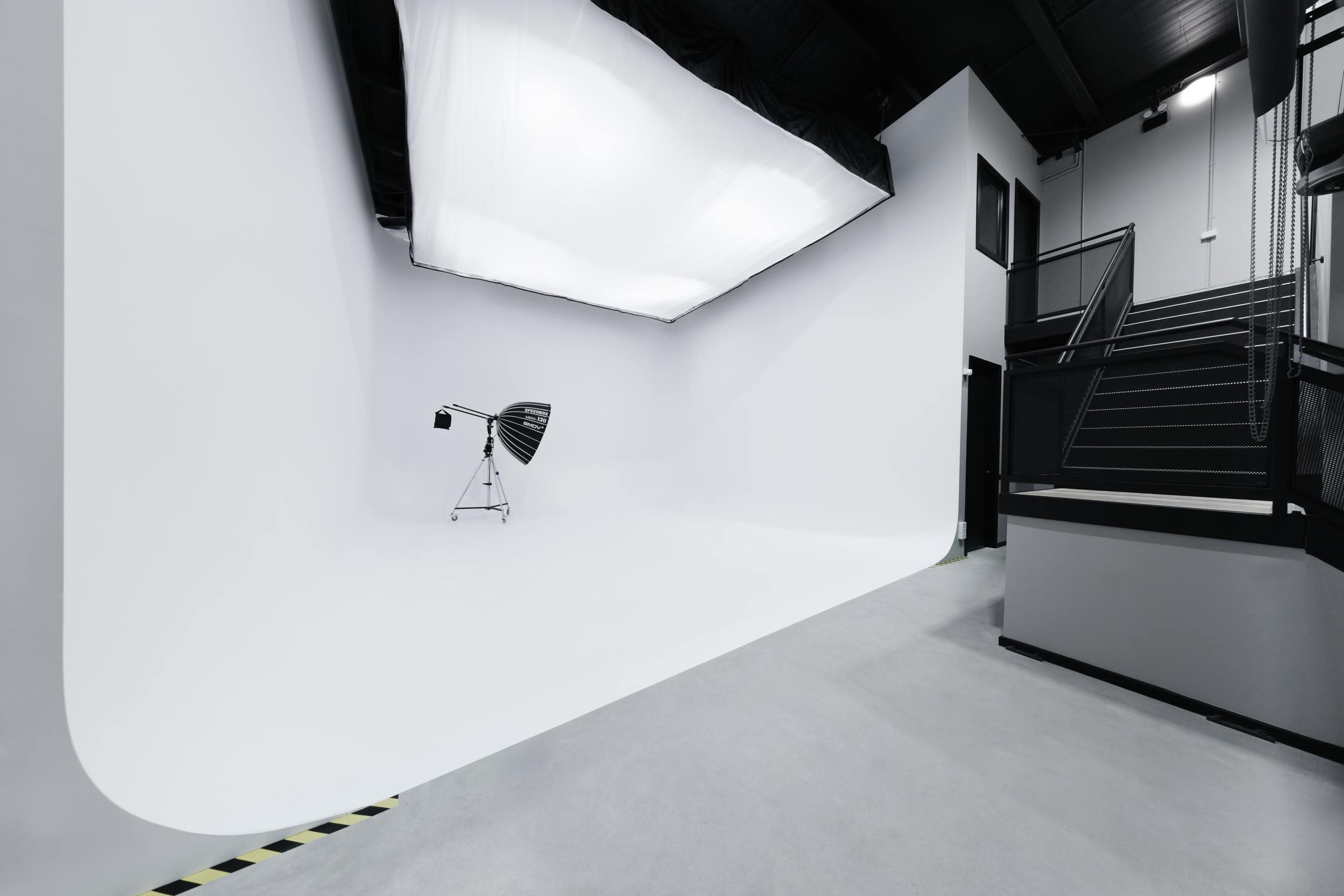The image shows a spacious photography studio with a seamless white backdrop, a light source on a stand, and a staircase leading to an upper level.