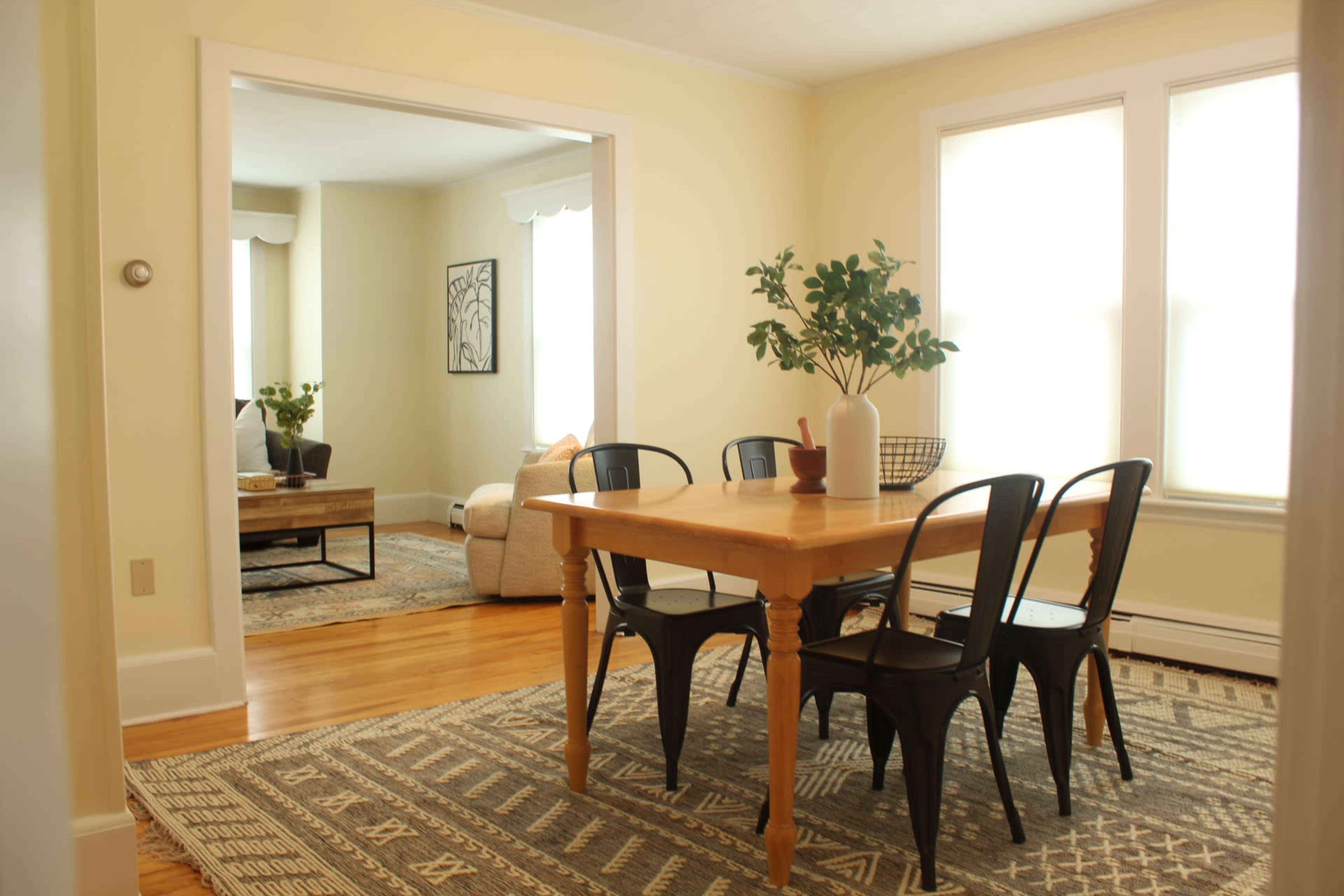 A dining area features a wooden table surrounded by black chairs, with a view into a bright living room through an open archway.