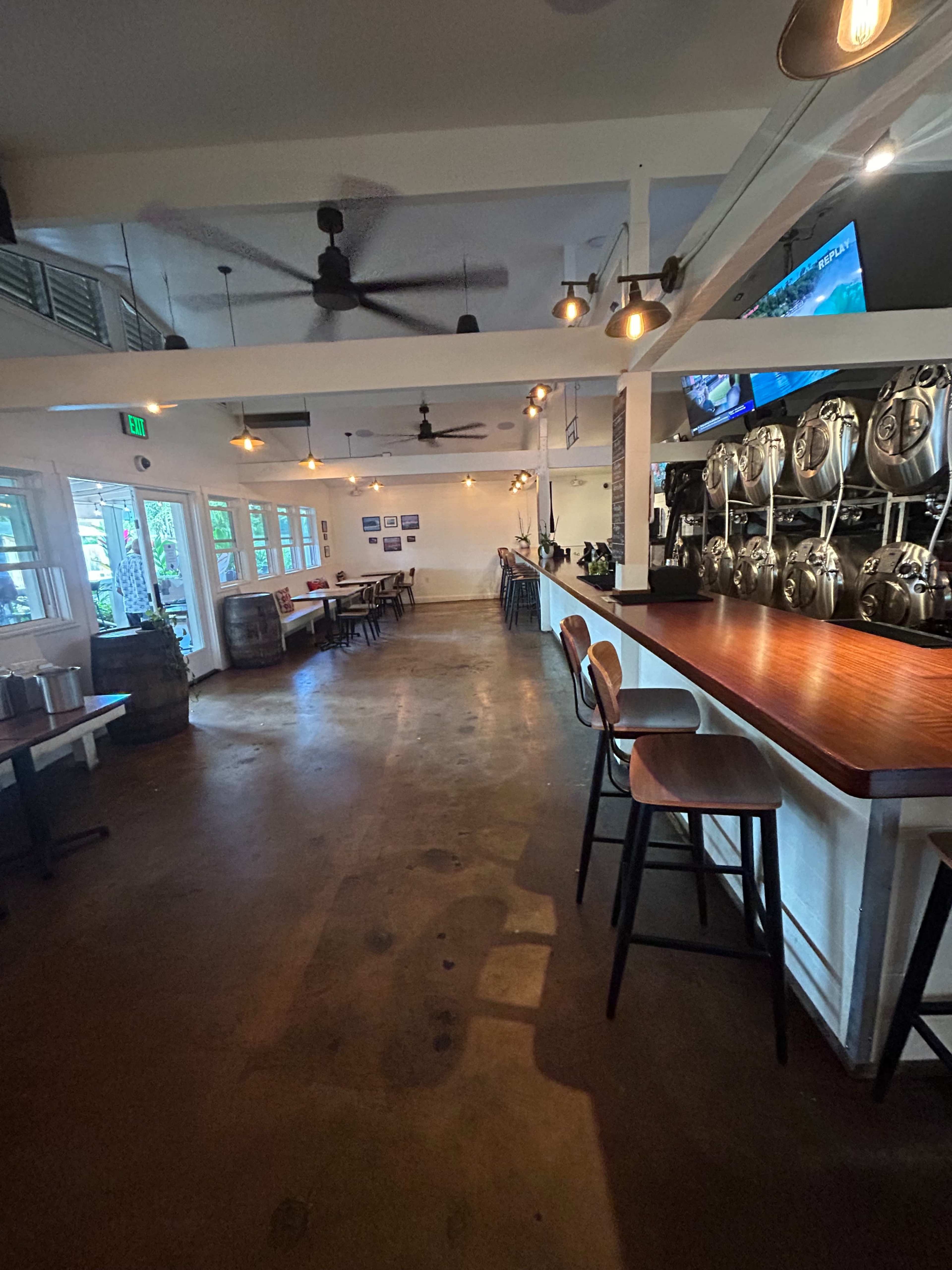 The image shows a spacious bar area with a long wooden counter, high stools, and metal beer kegs lined up against the wall, illuminated by pendant lights.