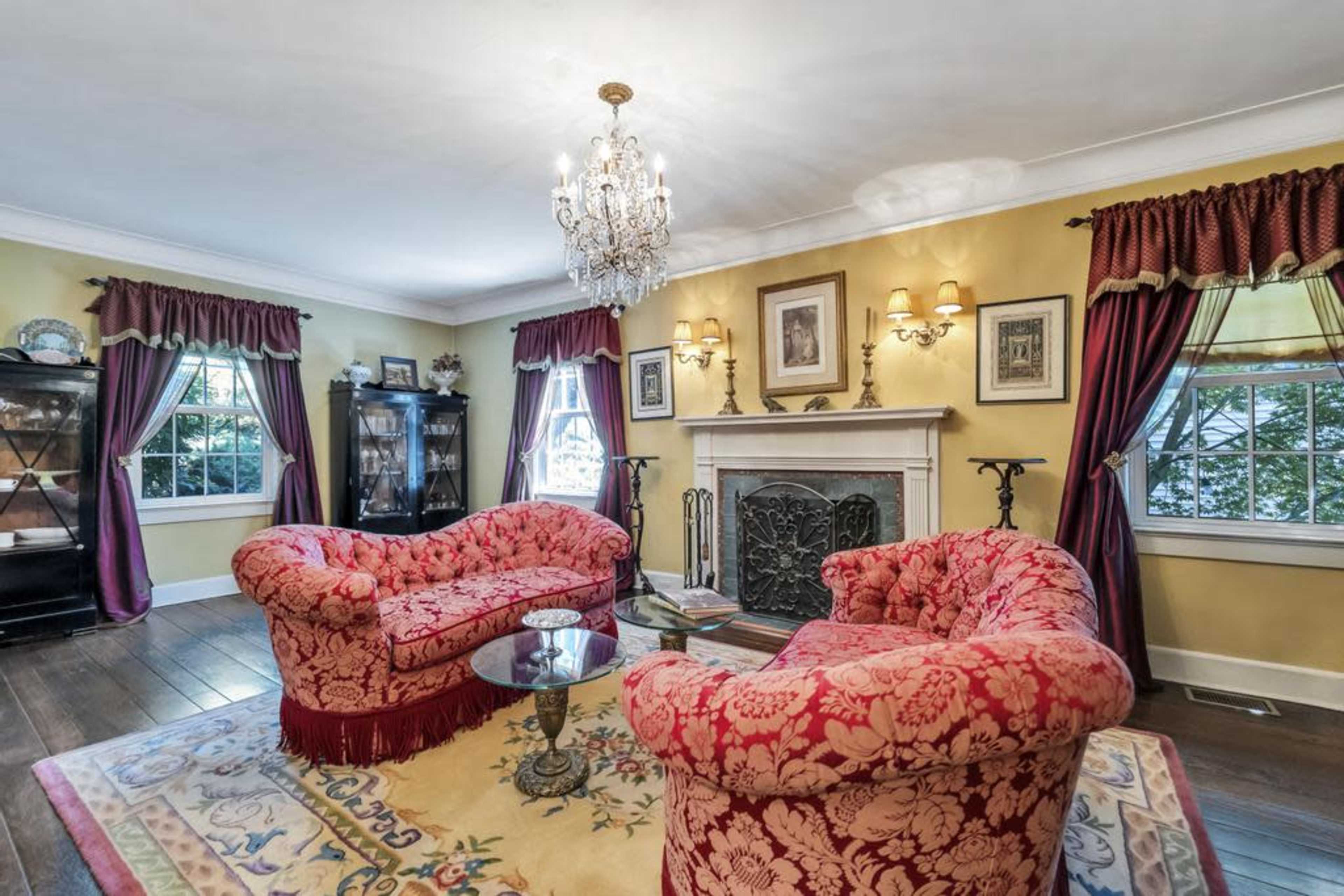A living room featuring two red upholstered sofas, a glass coffee table, a chandelier, and a fireplace, with yellow walls and large windows adorned with dark curtains.
