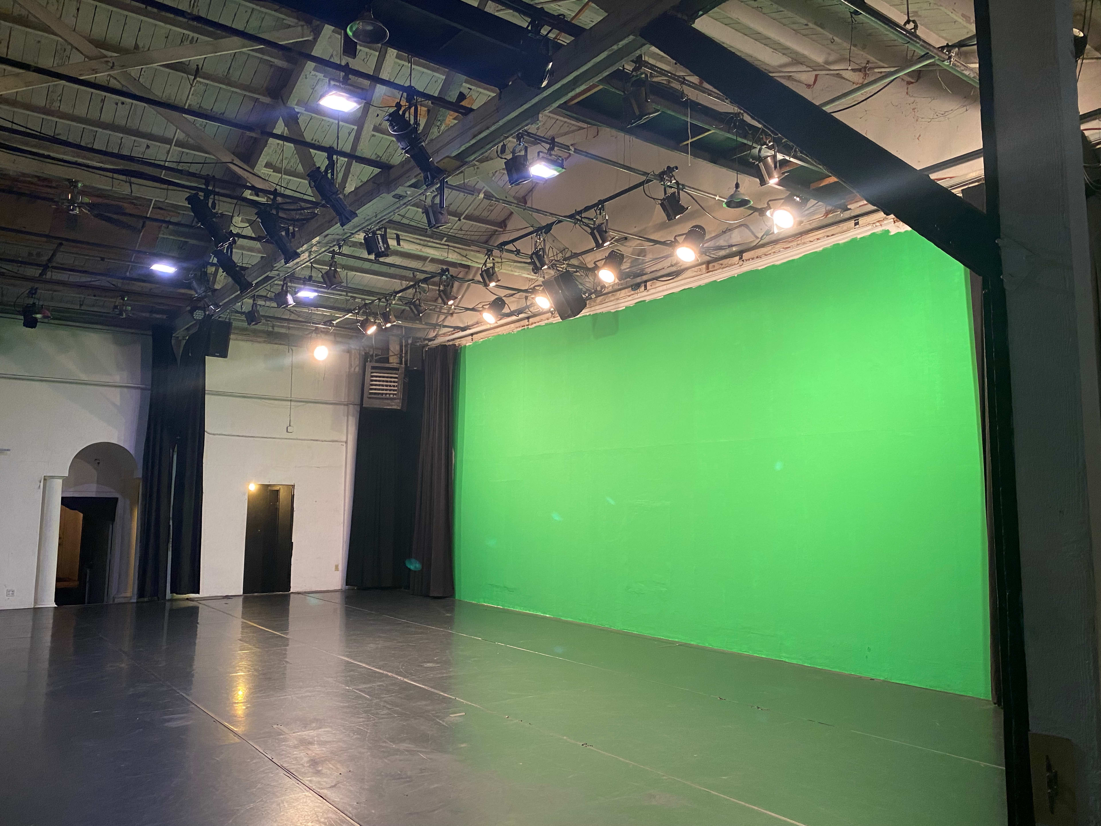 The image shows an empty studio space with a bright green screen on one wall and overhead lighting fixtures.