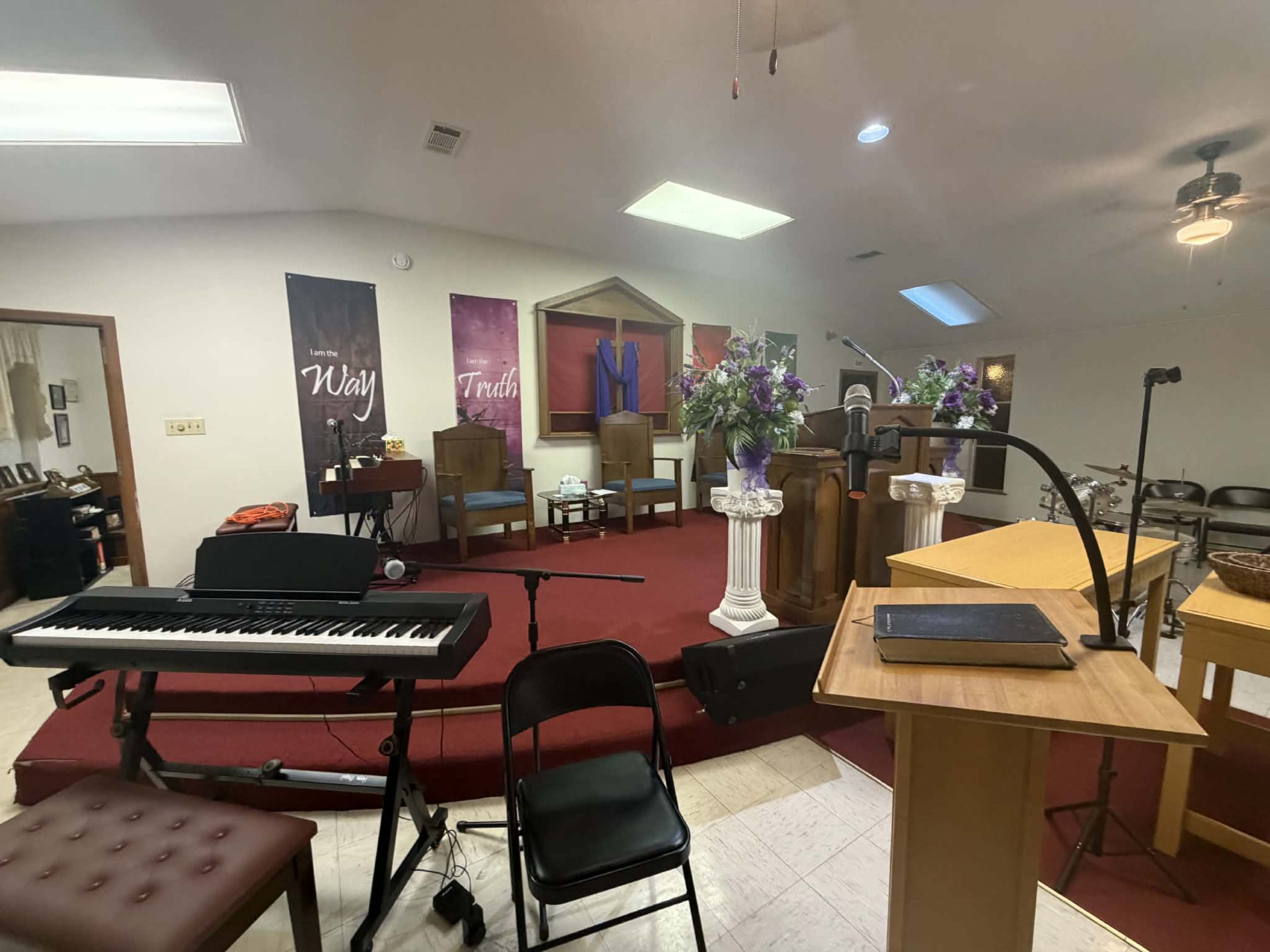 The image shows the interior of a small church, featuring a podium, a keyboard, and decorative banners along the walls, while floral arrangements are placed on the altar.