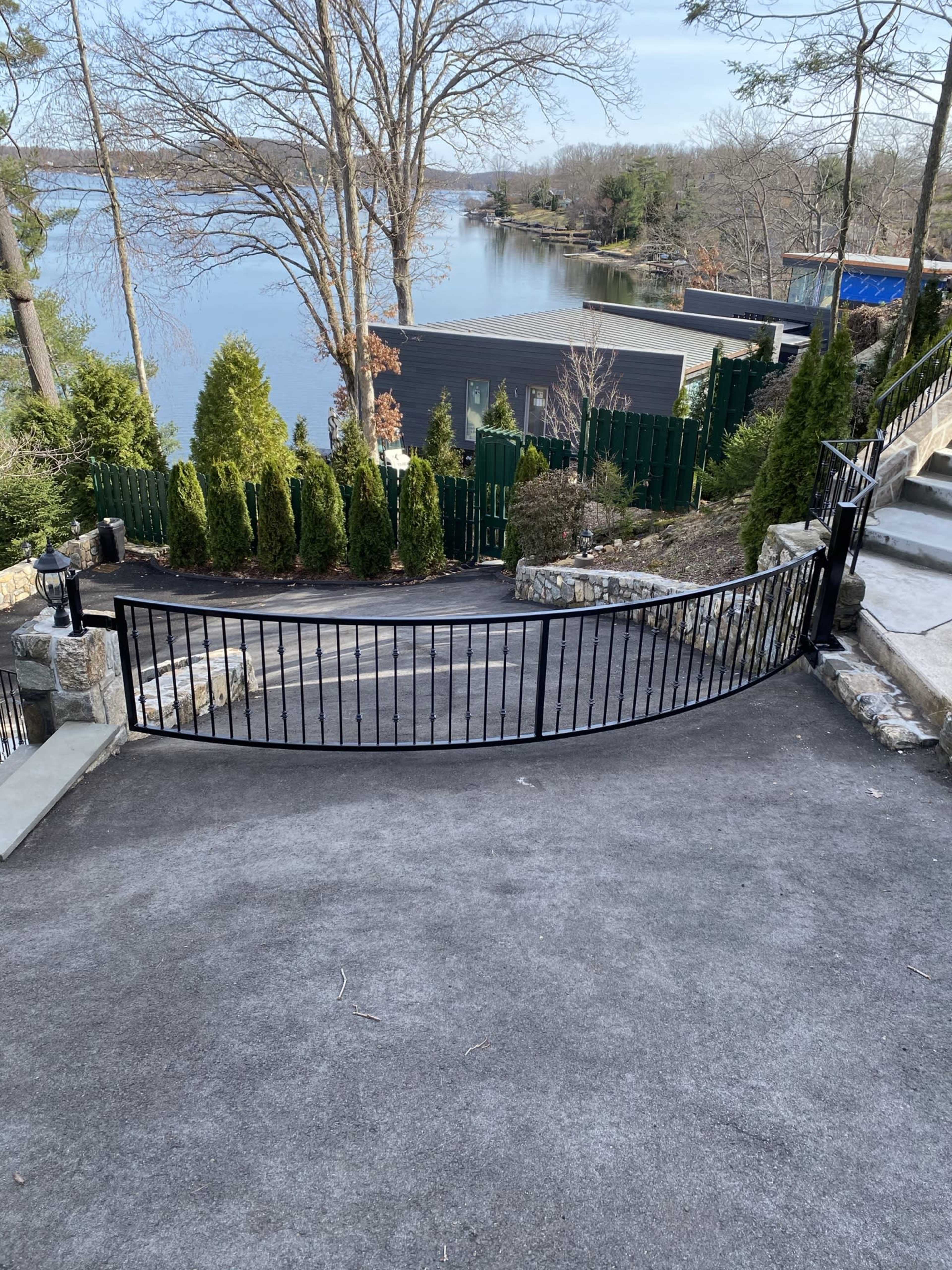 A curved black gate leads to a gravel path overlooking a calm lake, bordered by neatly trimmed trees and shrubs.