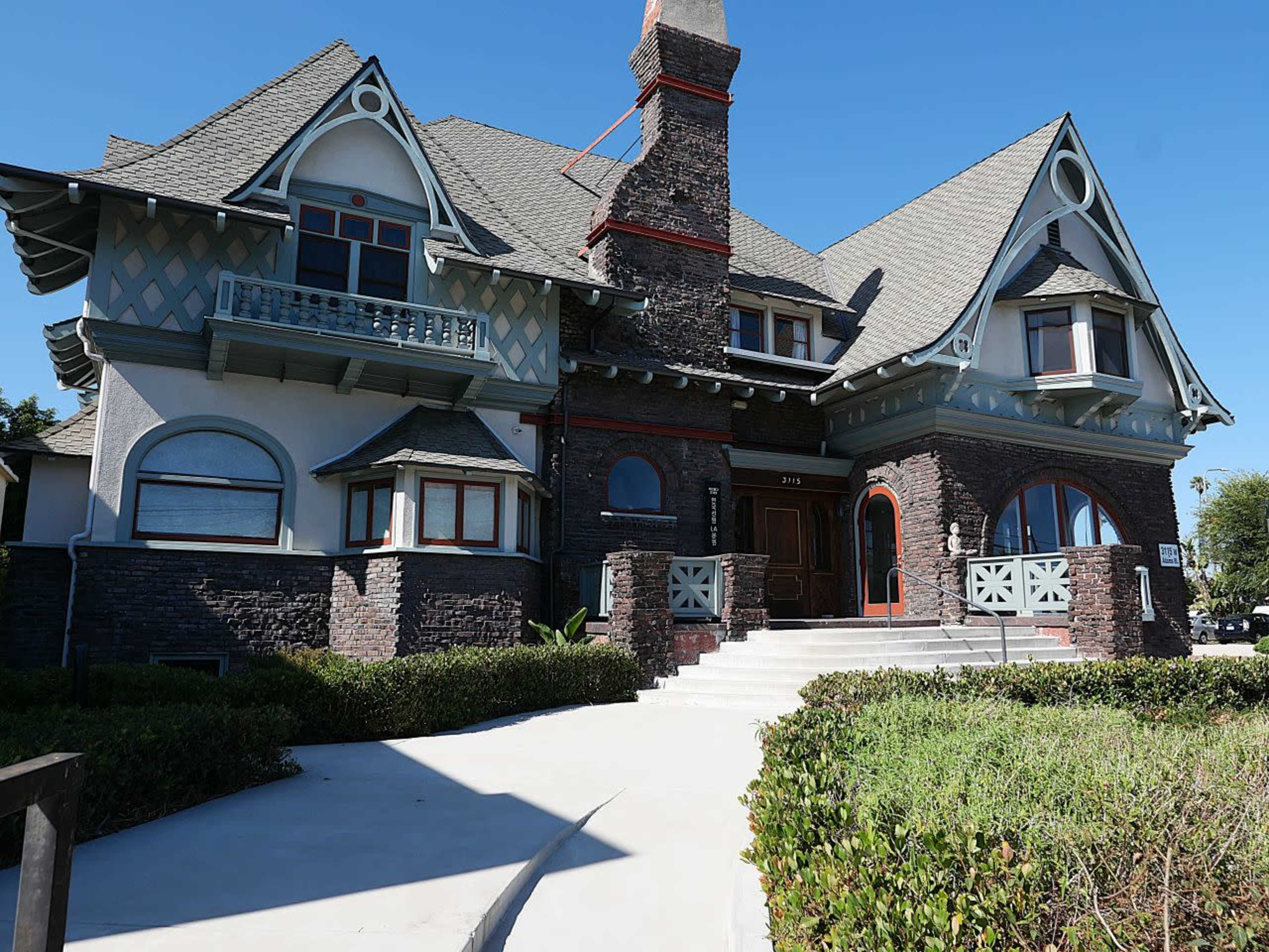 The image shows a large, ornate house with a mix of stone and wood features, intricate rooflines, and a well-maintained front pathway leading up to the entrance.