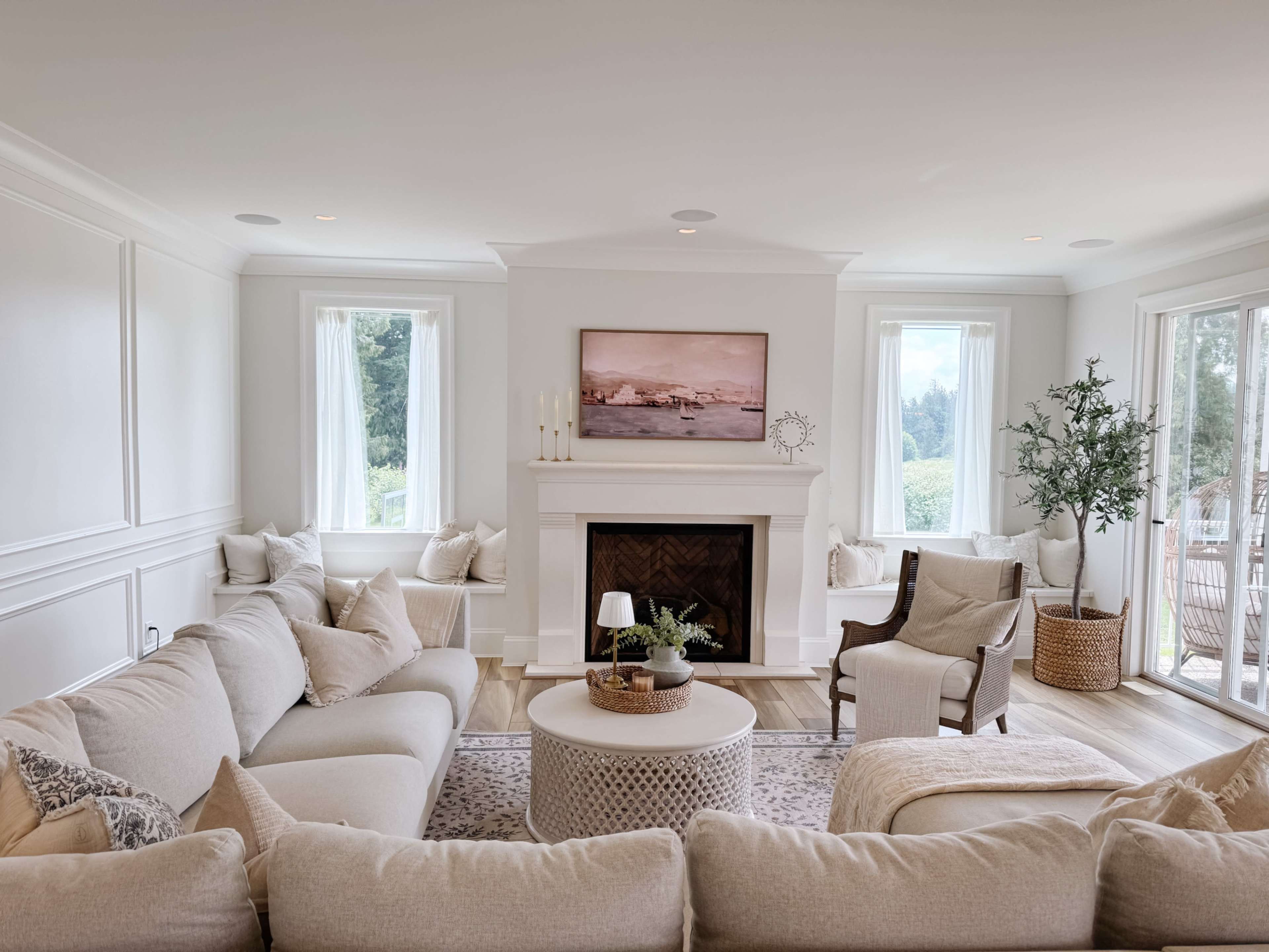 The room features a light-filled living area with a white fireplace, large windows, a circular coffee table, and a mix of comfortable seating, including a sofa and an armchair.