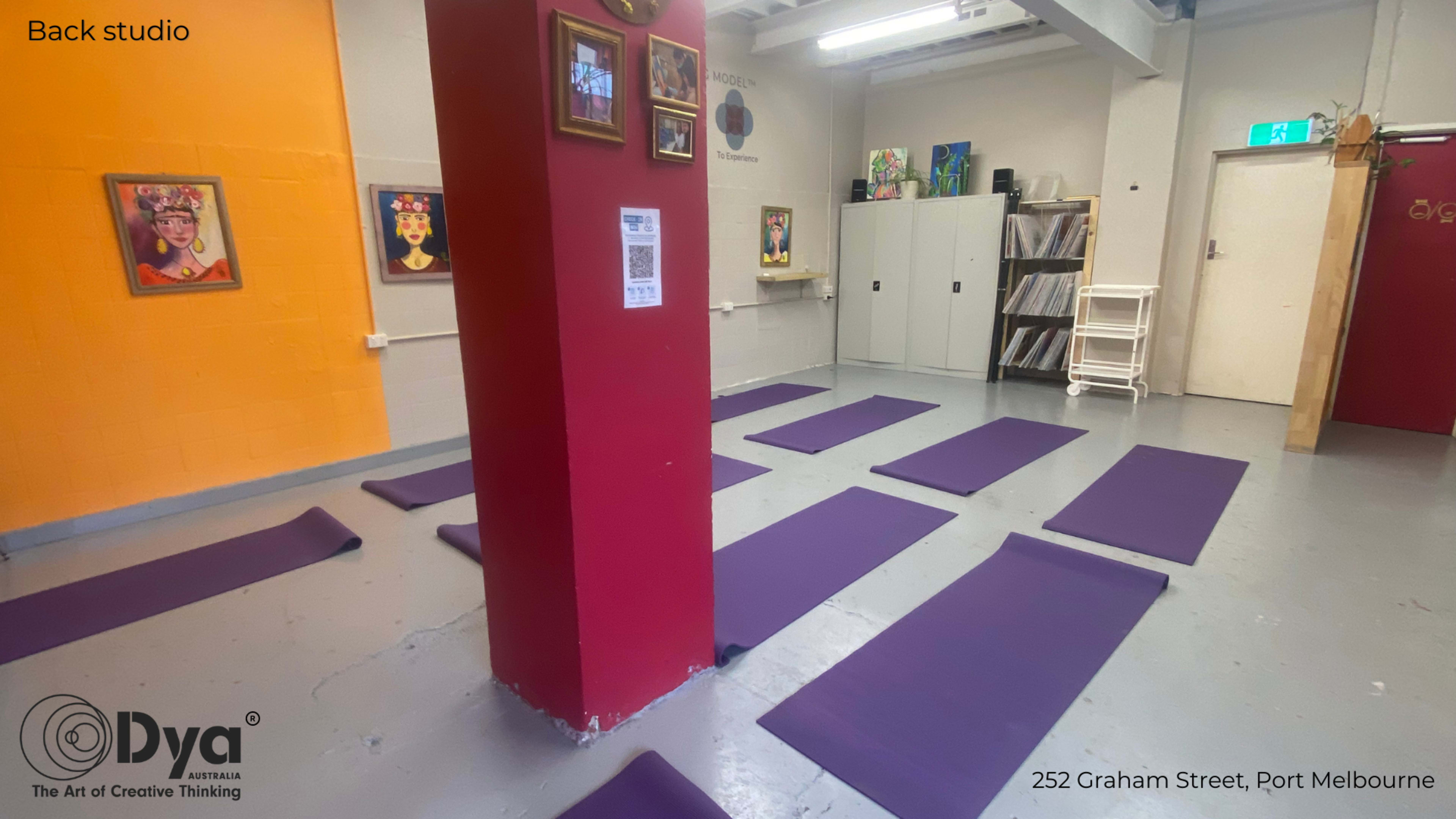 A back studio with multiple purple yoga mats neatly arranged on the floor, surrounded by walls featuring artwork and a storage cabinet.