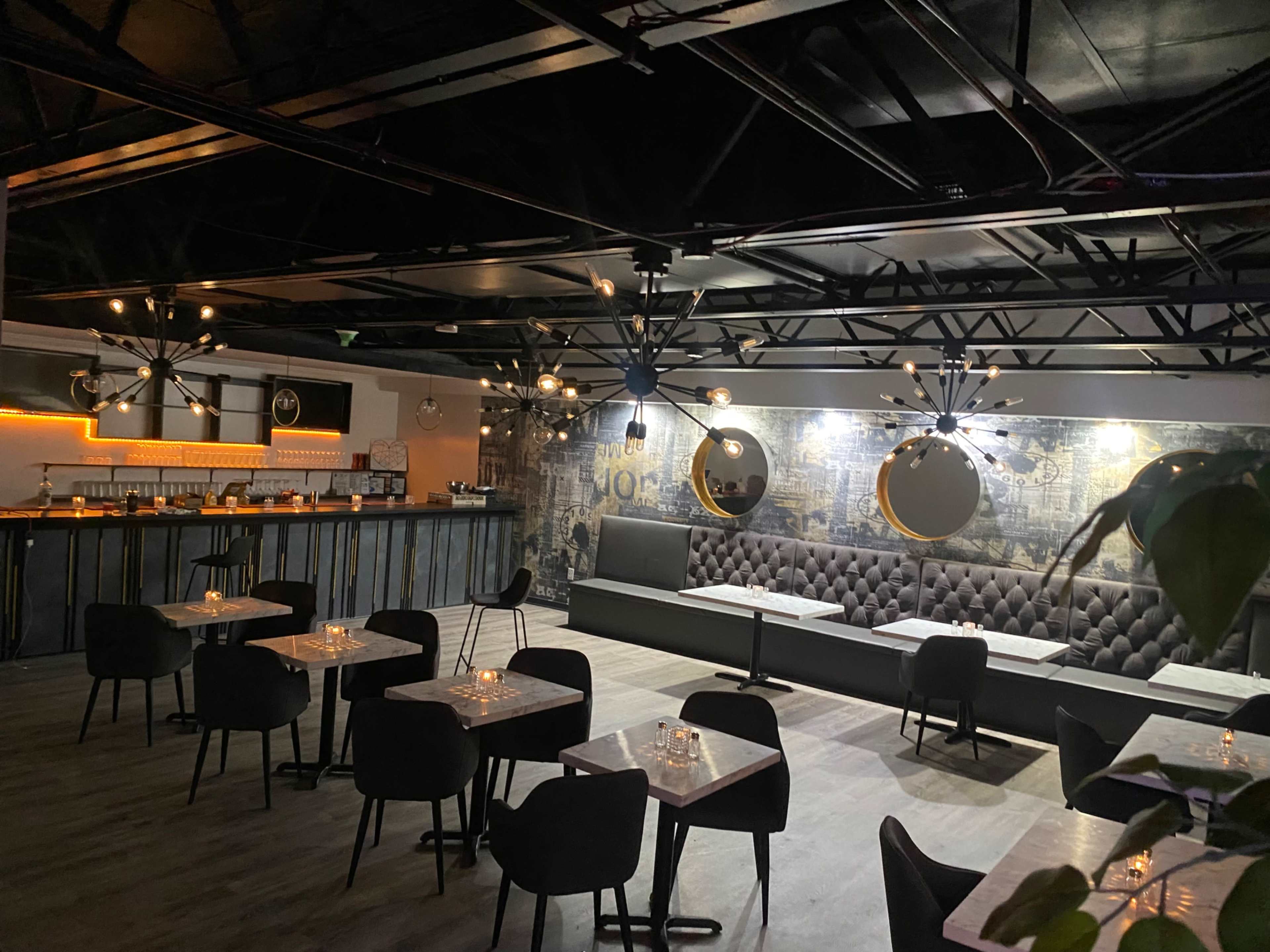 The image shows a modern restaurant interior featuring a bar along one wall, several tables with candles, and a plush seating area with decorative lighting.