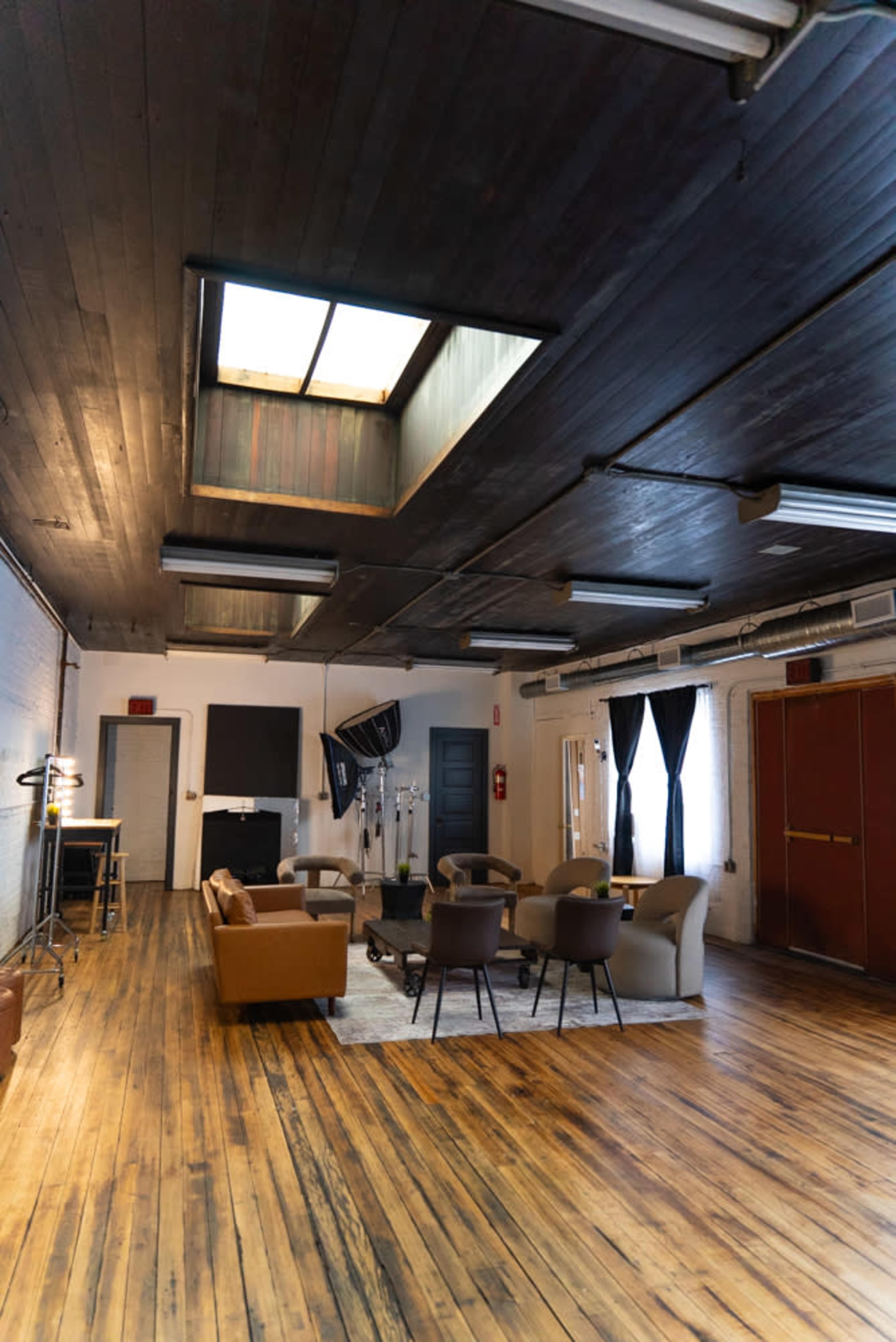The image shows a spacious, well-lit room with wooden flooring, featuring a seating area with couches and chairs, a skylight, and various lighting equipment.