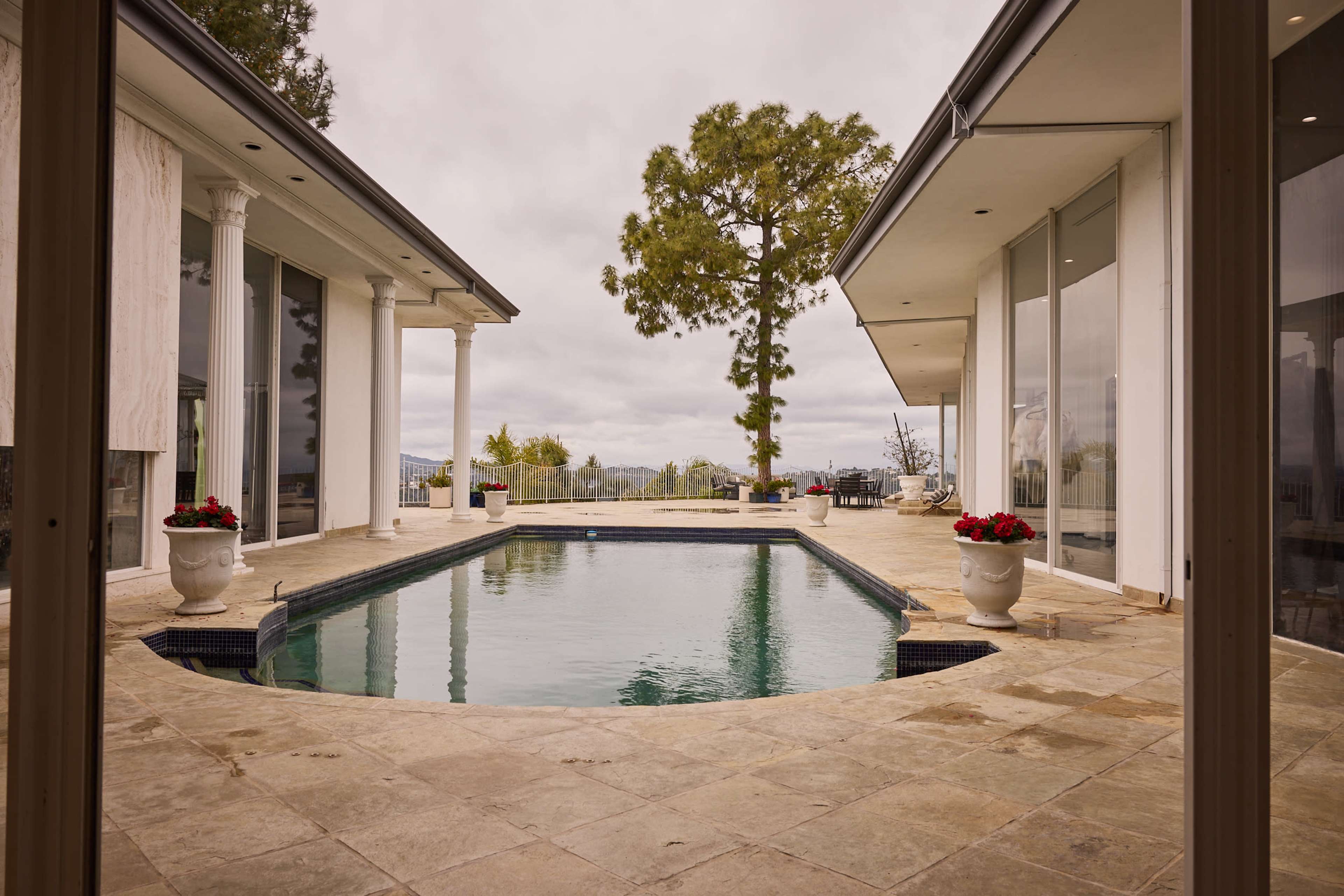 A spacious modern home features an infinity pool surrounded by stone tiles and large windows with views of distant hills and trees.