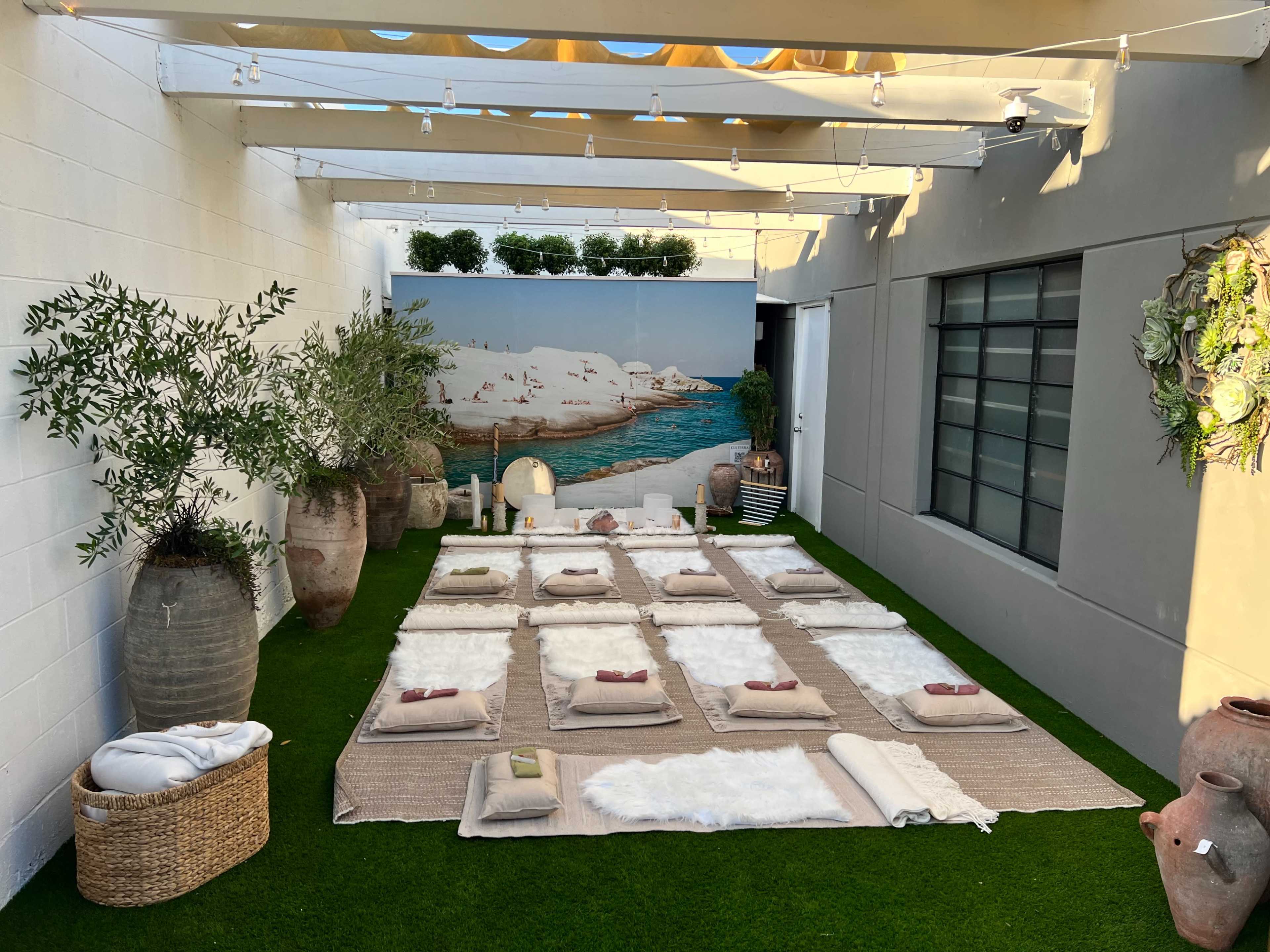 The scene shows a tranquil outdoor space set up for relaxation, featuring neatly arranged mats and cushions on a grassy floor under a pergola with string lights.