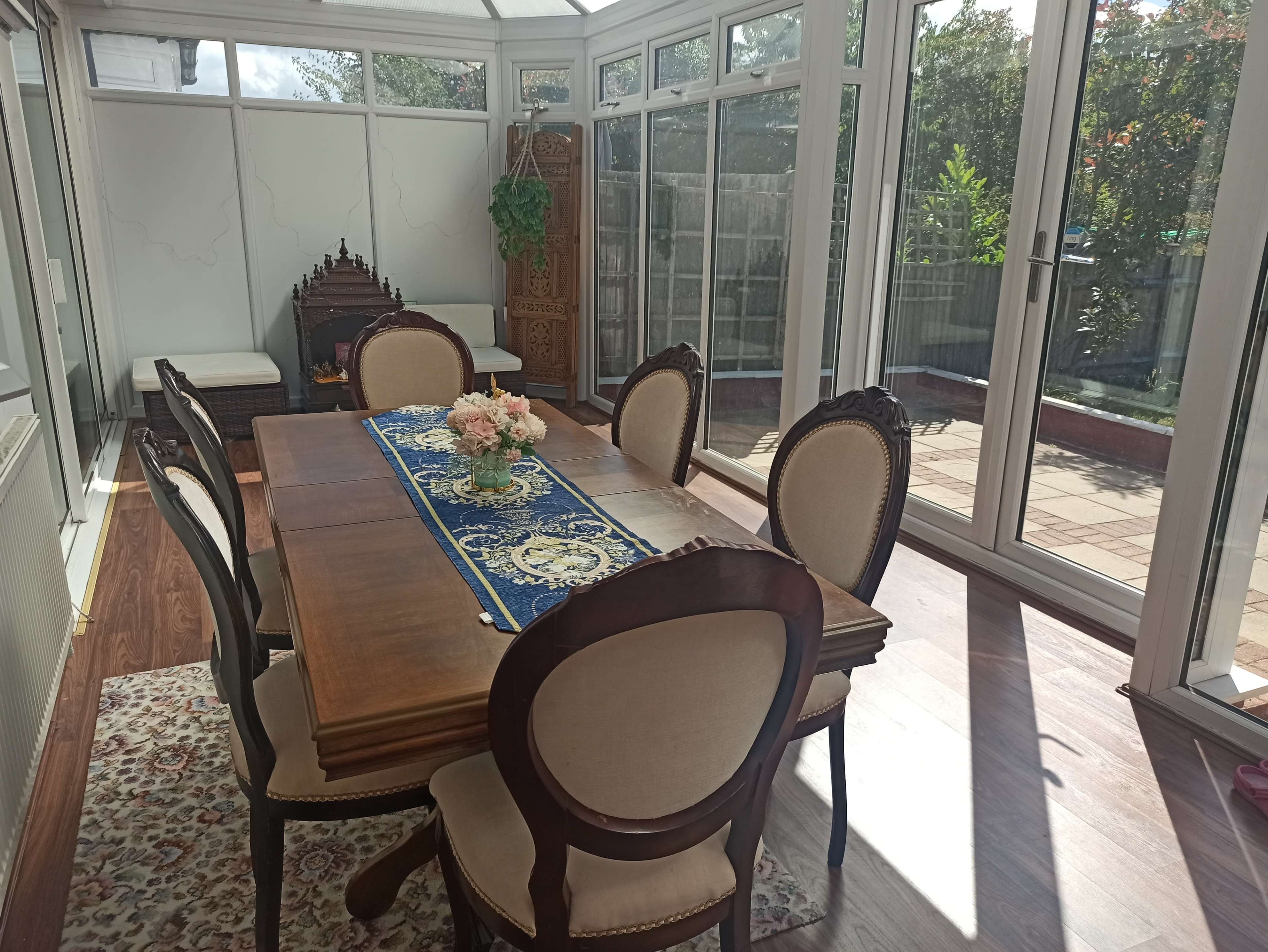 A wooden dining table with upholstered chairs is set in a sunlit conservatory, accented by a decorative runner and a vase of flowers.