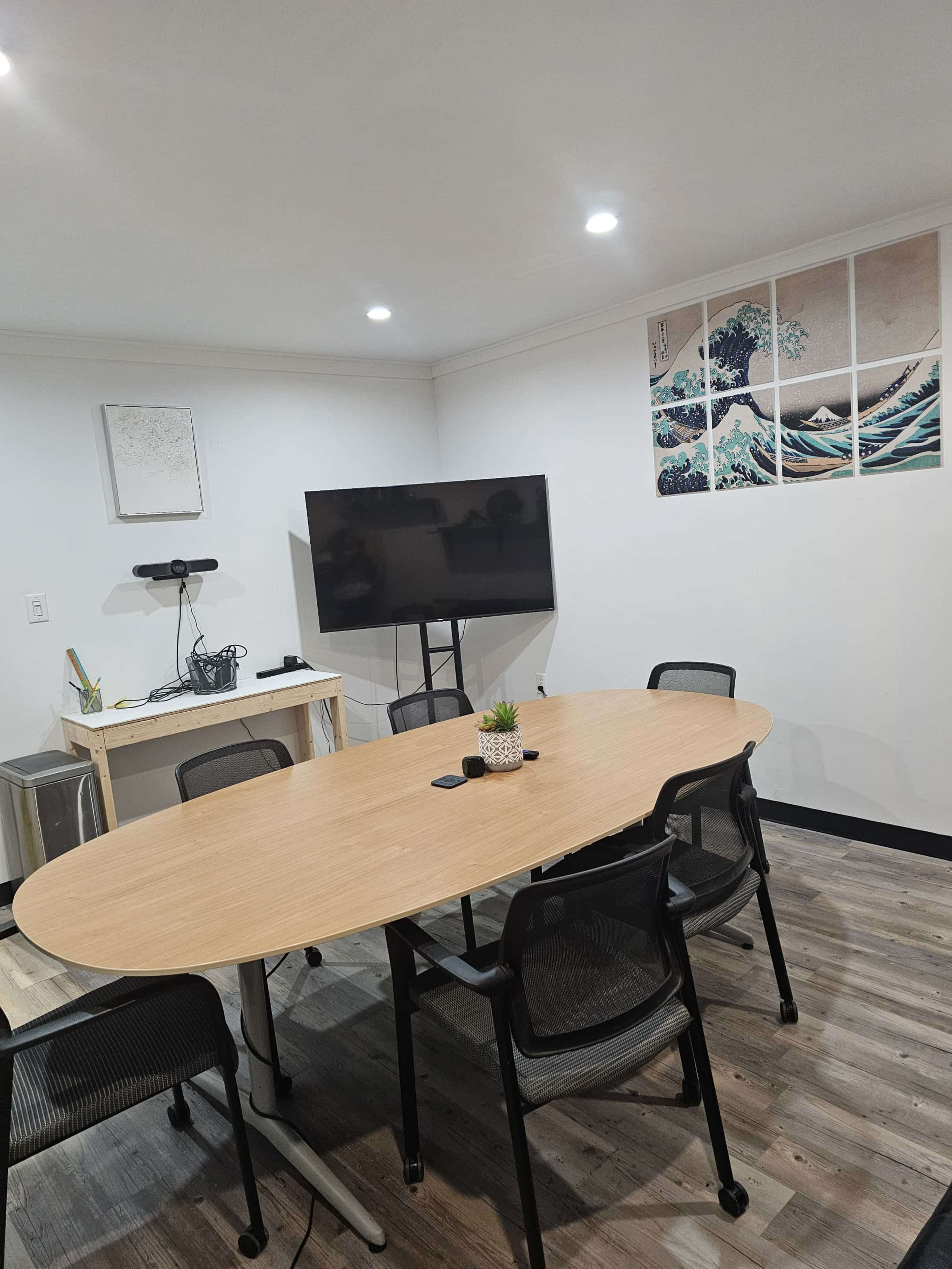 The image shows a modern conference room featuring a large wooden table surrounded by black chairs, a television mounted on a stand, and a wall decorated with artwork.
