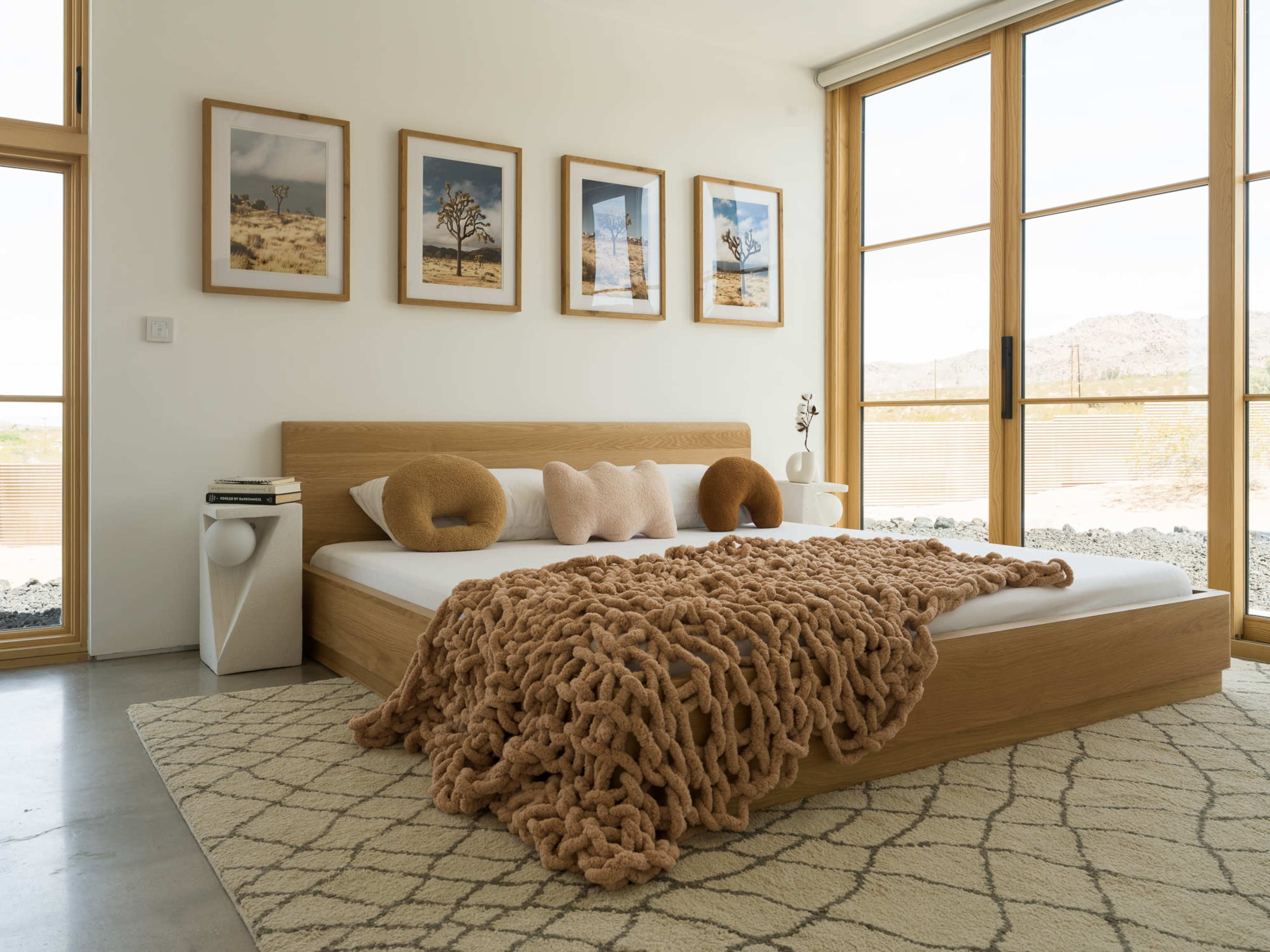A modern bedroom features a wooden bed with a large knitted blanket, framed photographs on the walls, and large windows allowing ample natural light.