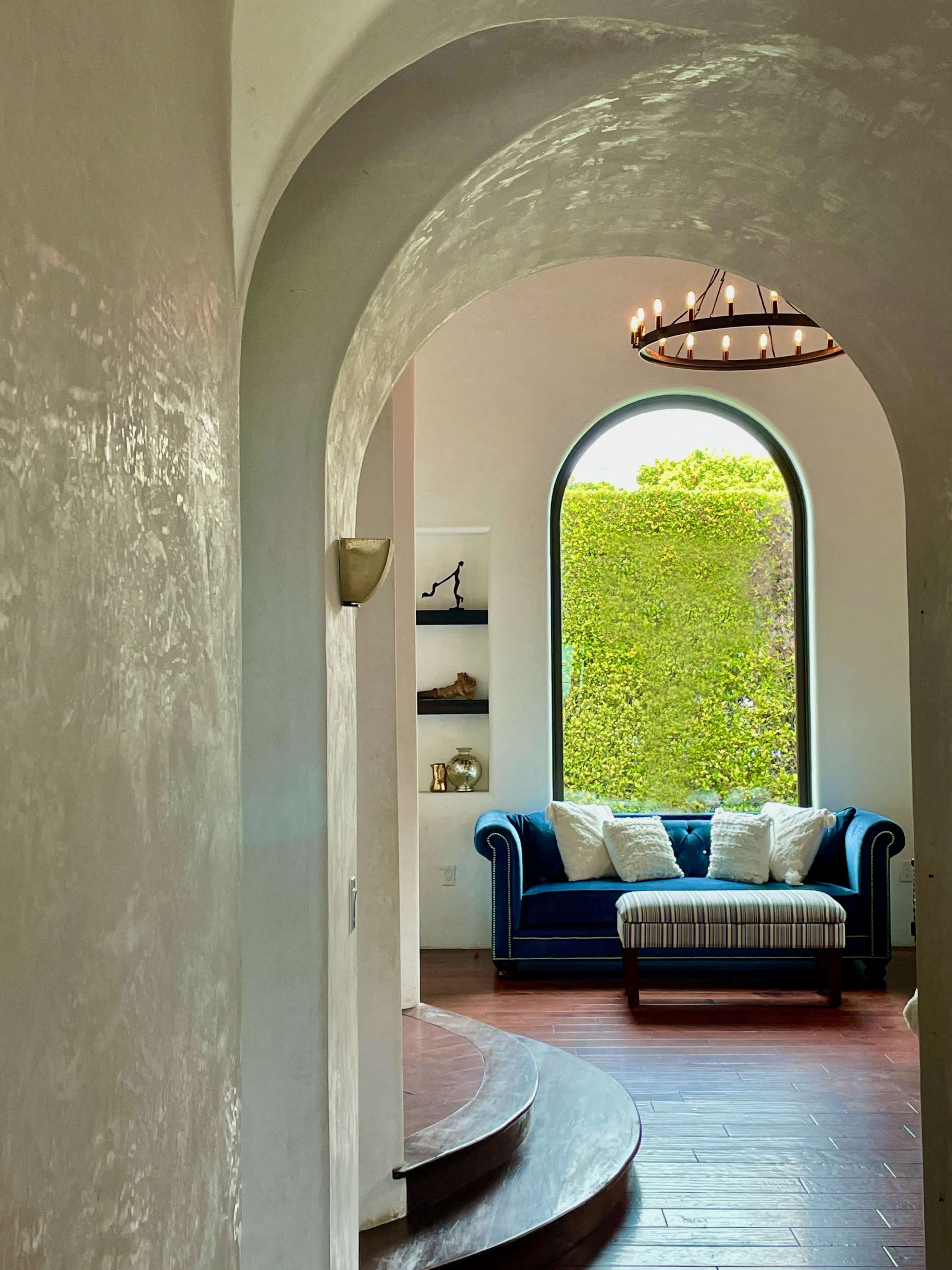 A hallway with a curved arch leading to a living area featuring a blue sofa and a large window with greenery outside.