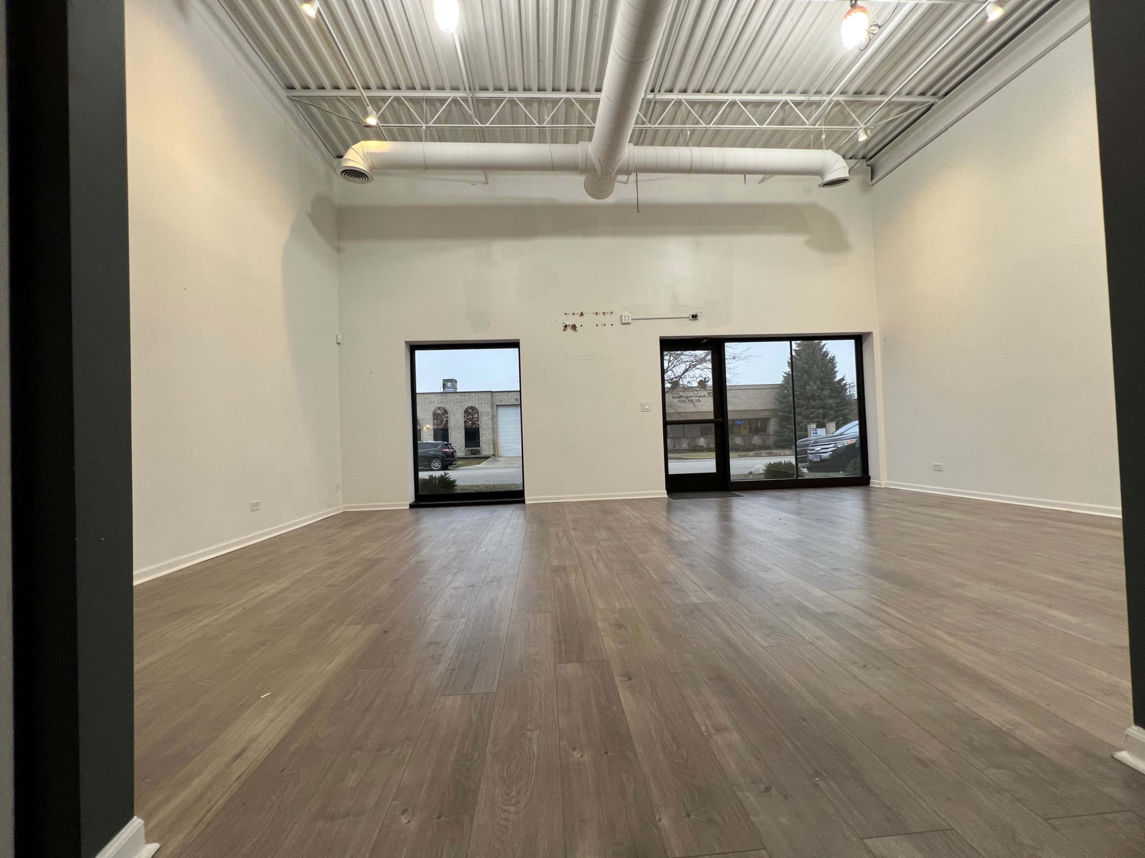 An empty room with wooden flooring and large windows along one wall, allowing natural light to enter.
