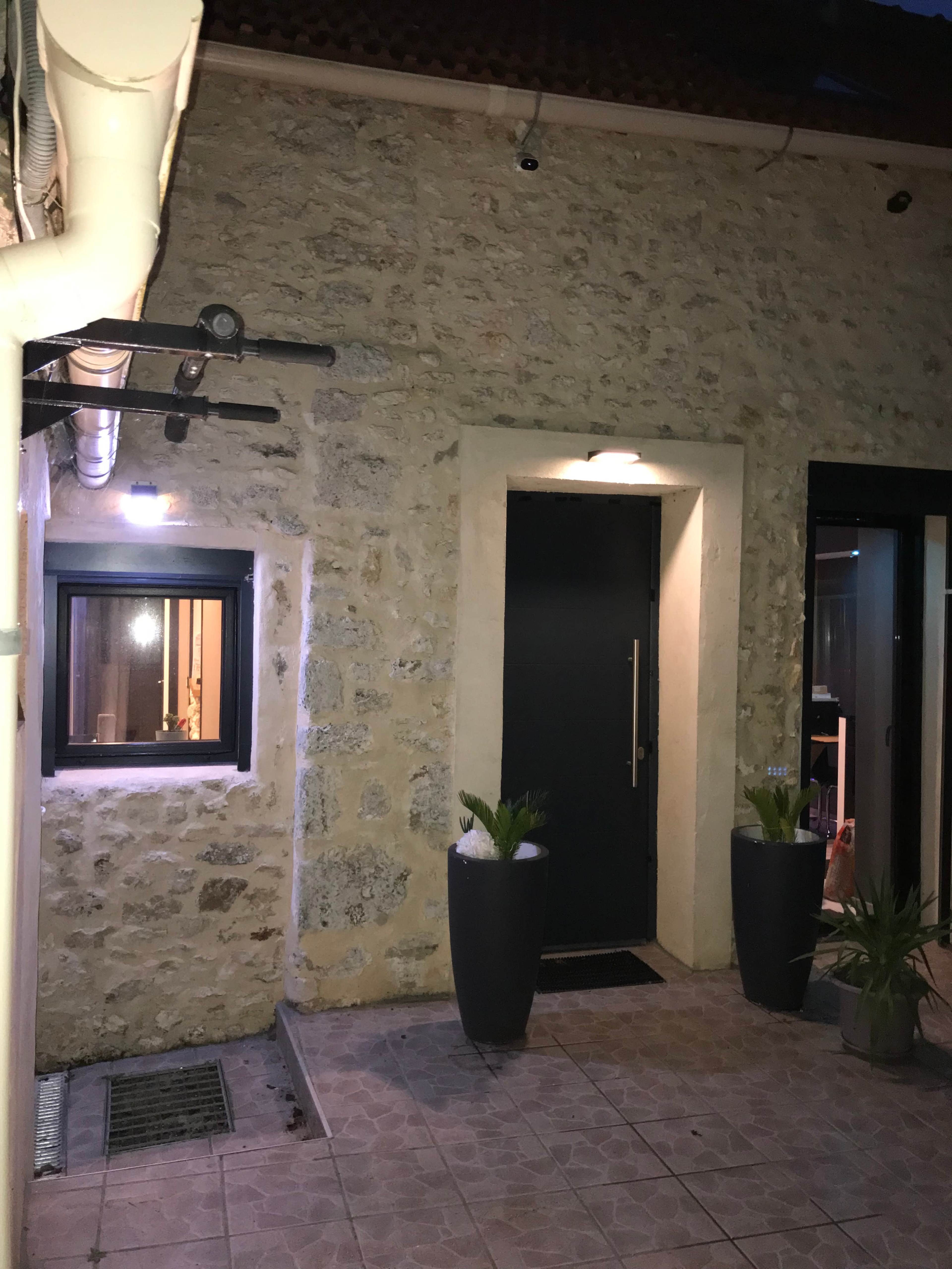 A stone wall building with a dark front door, a small window, and two potted plants near the entrance, illuminated by exterior lights.