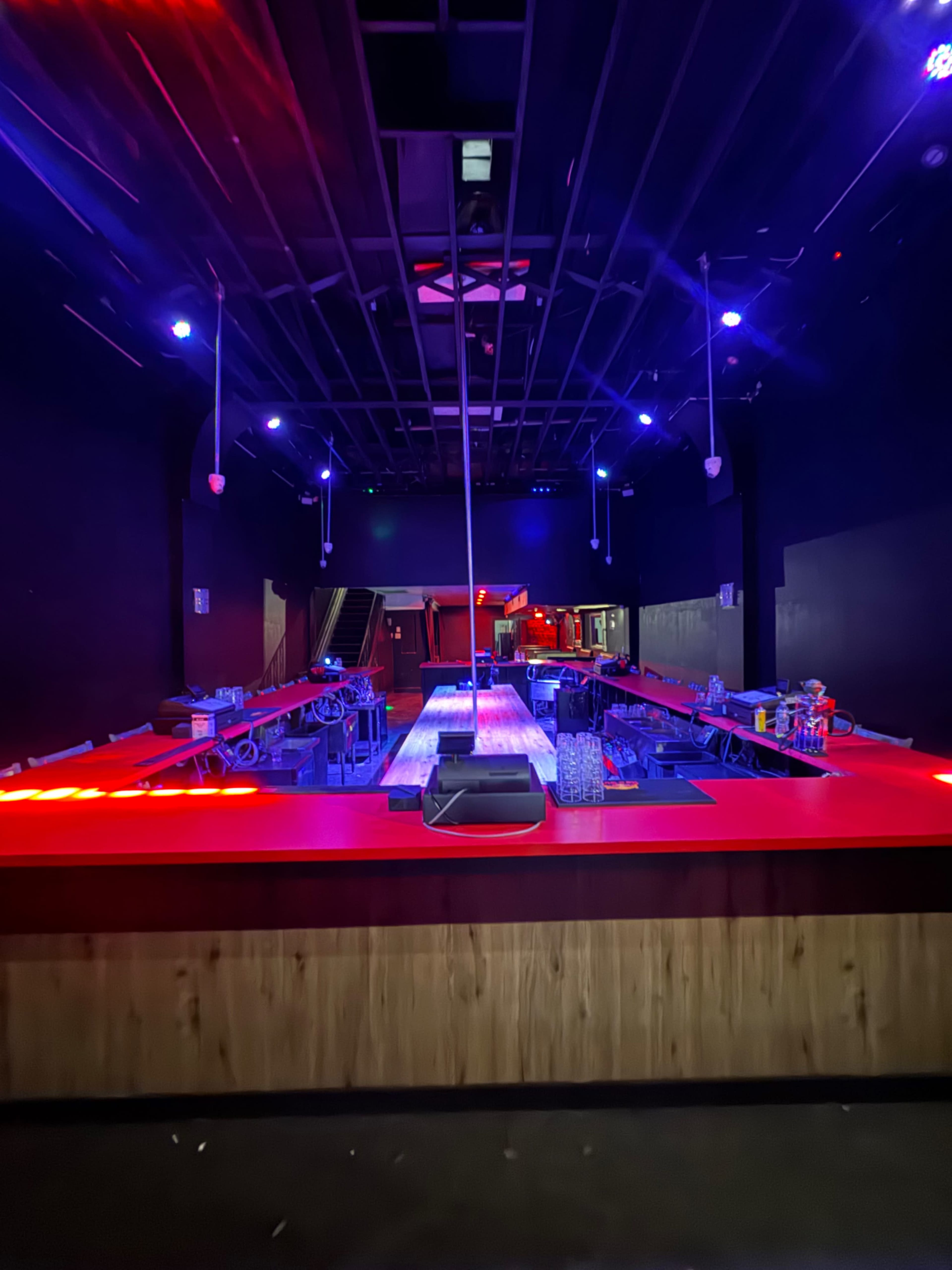 The image shows a dimly lit bar area with a long counter, illuminated by colorful lights and featuring empty tables and a pole in the center.