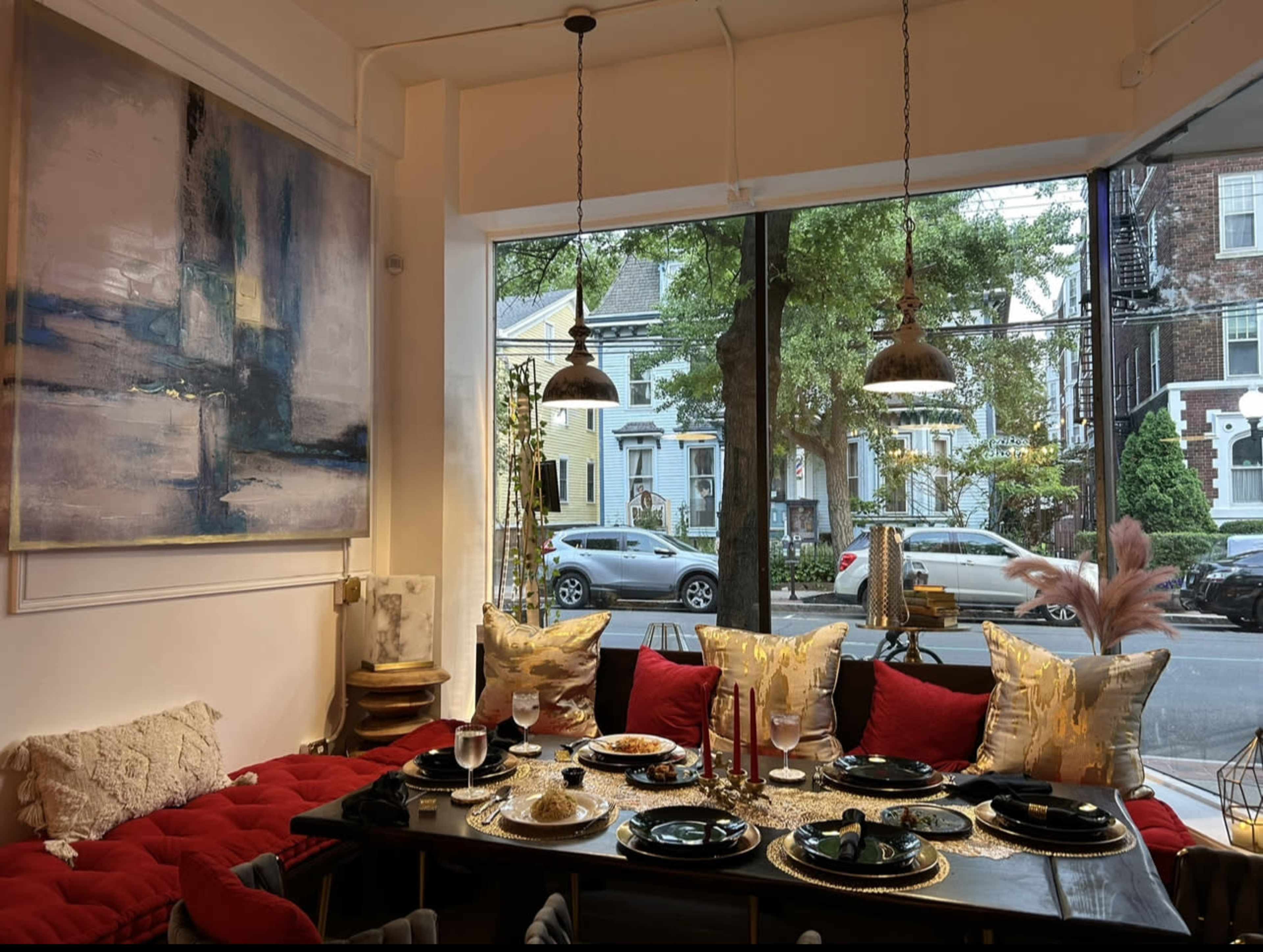 A dining area features a table set with elegant dishes, surrounded by cushions and adorned with artistic lighting, overlooking a street lined with trees and houses.