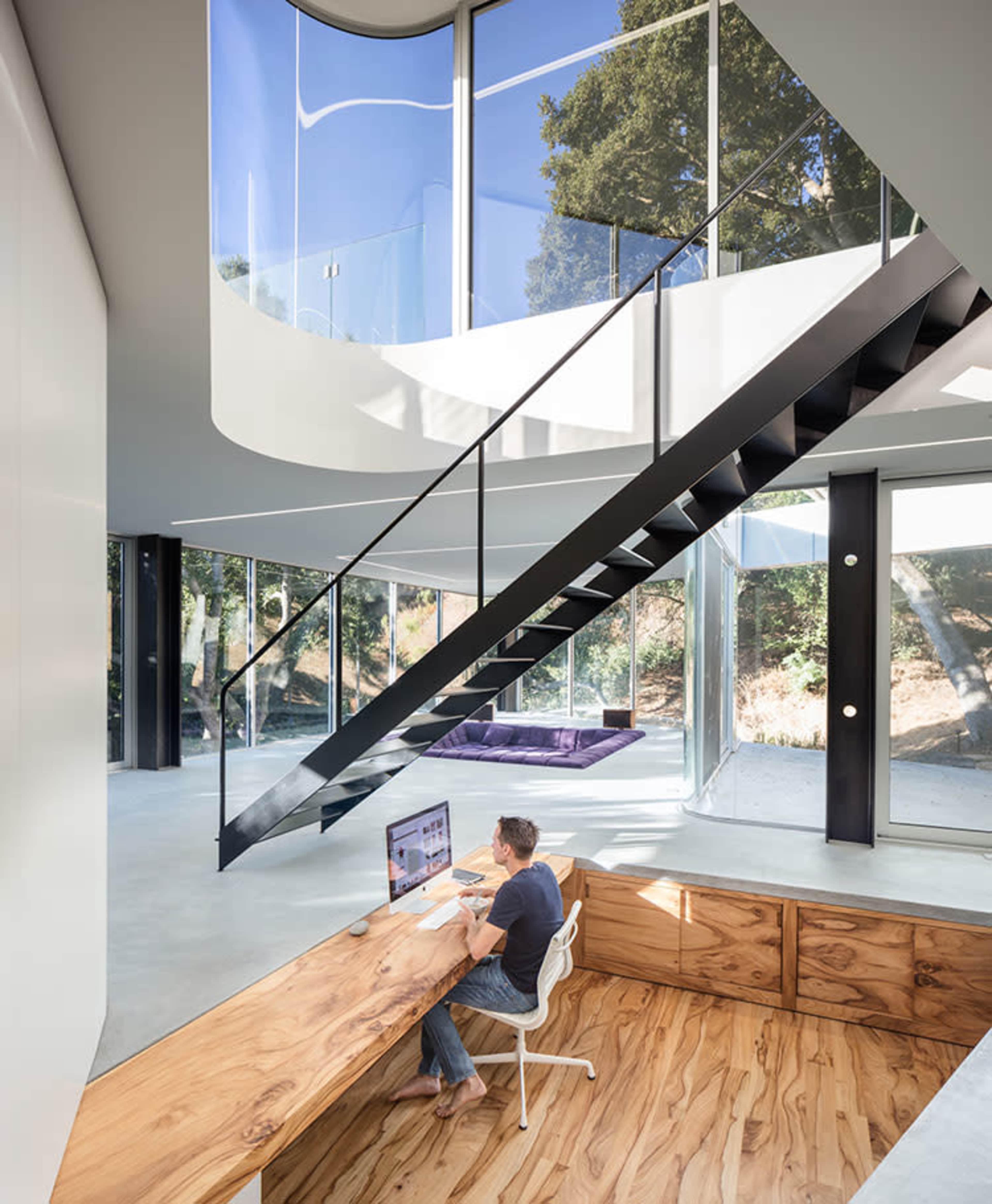 A person works at a minimalist wooden desk in a contemporary home with large windows and a spiral staircase.