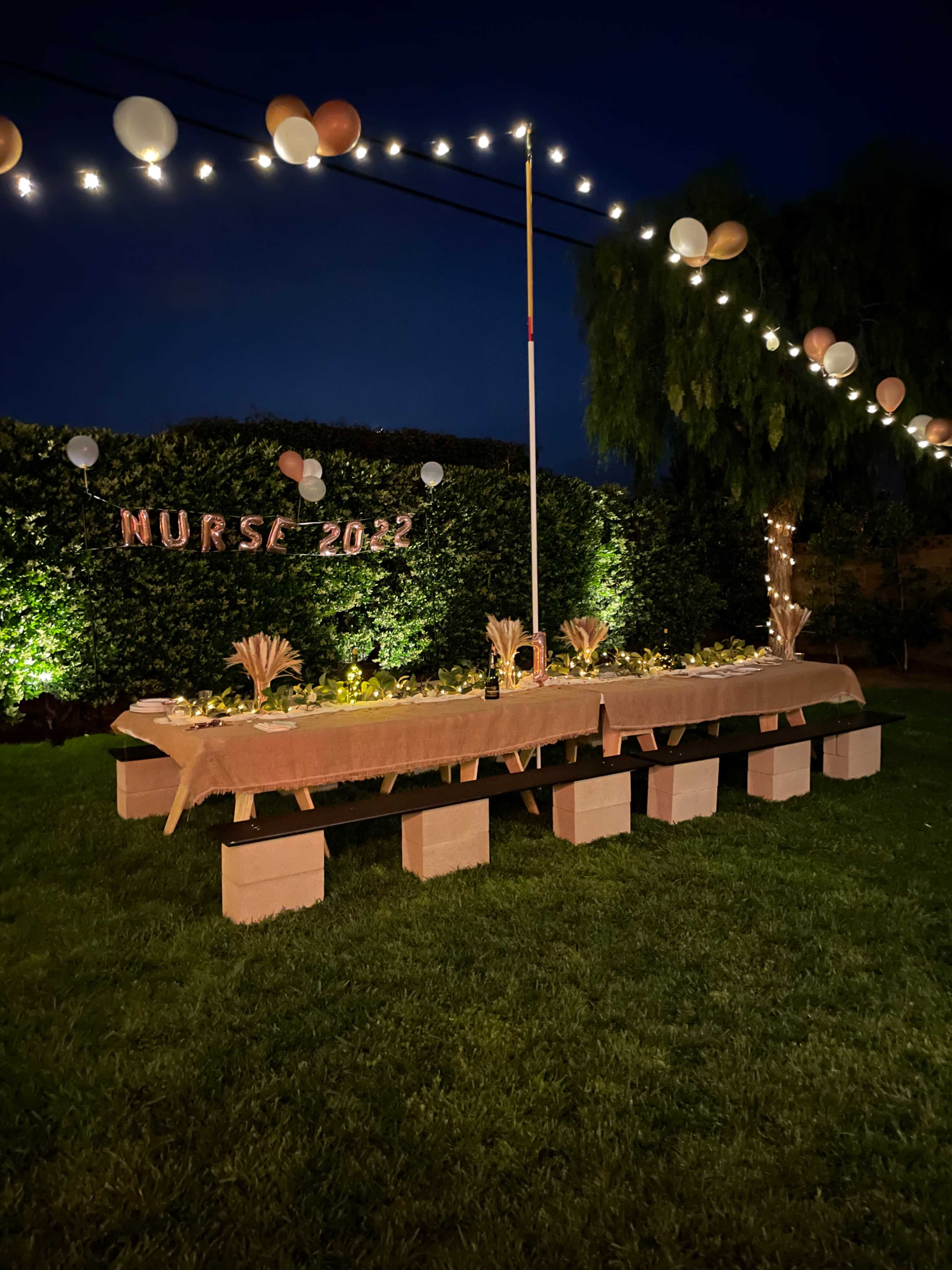 A long table is set with decorations and illuminated by string lights in a garden area for a celebration labeled "NURSE 2022."