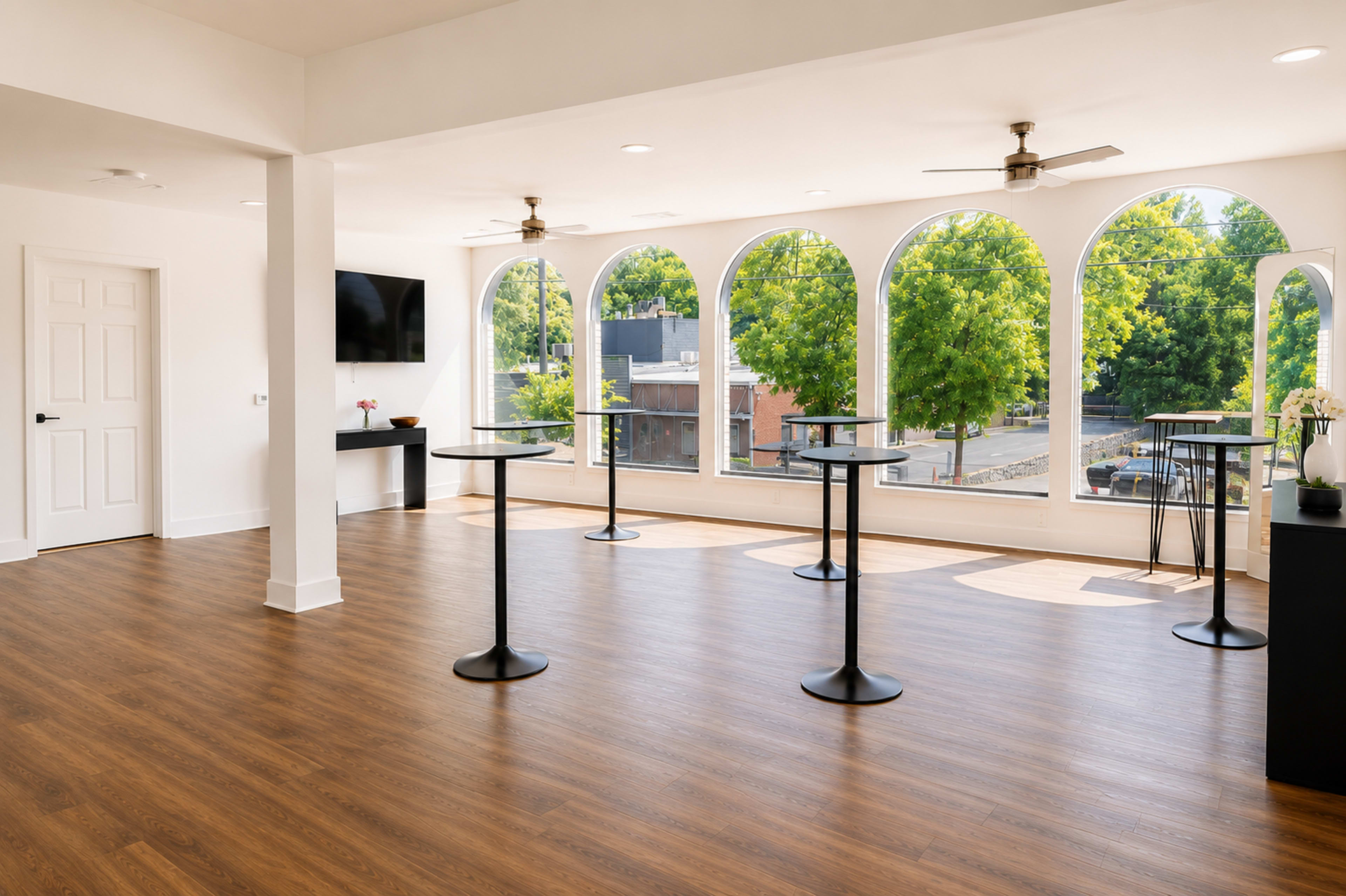 The image shows a spacious room with large arched windows, wooden flooring, and a few tall, round tables set up for an event.