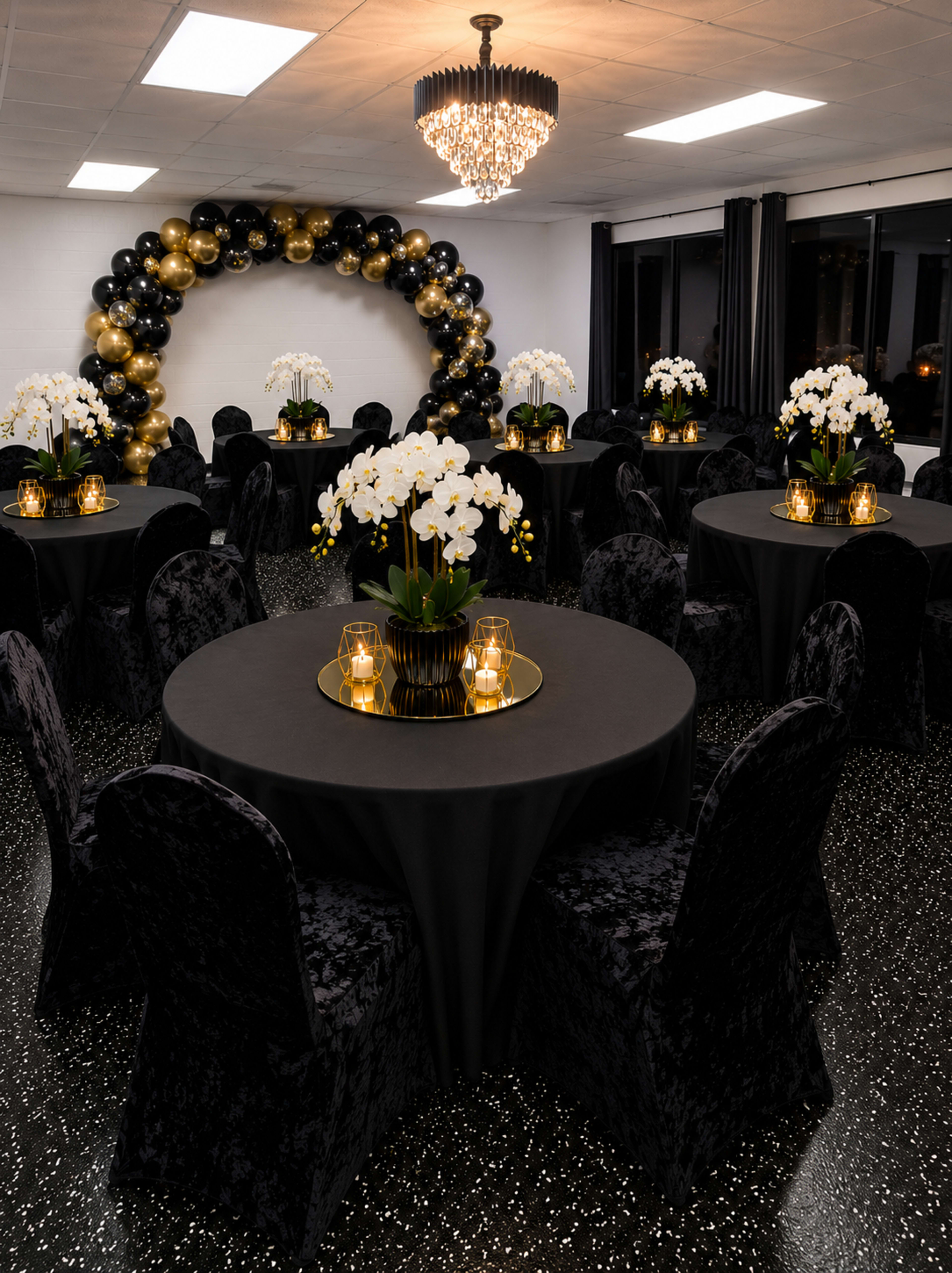 A banquet hall features round black tables adorned with elegant floral centerpieces, lit candles, and a backdrop of a black and gold balloon arch.