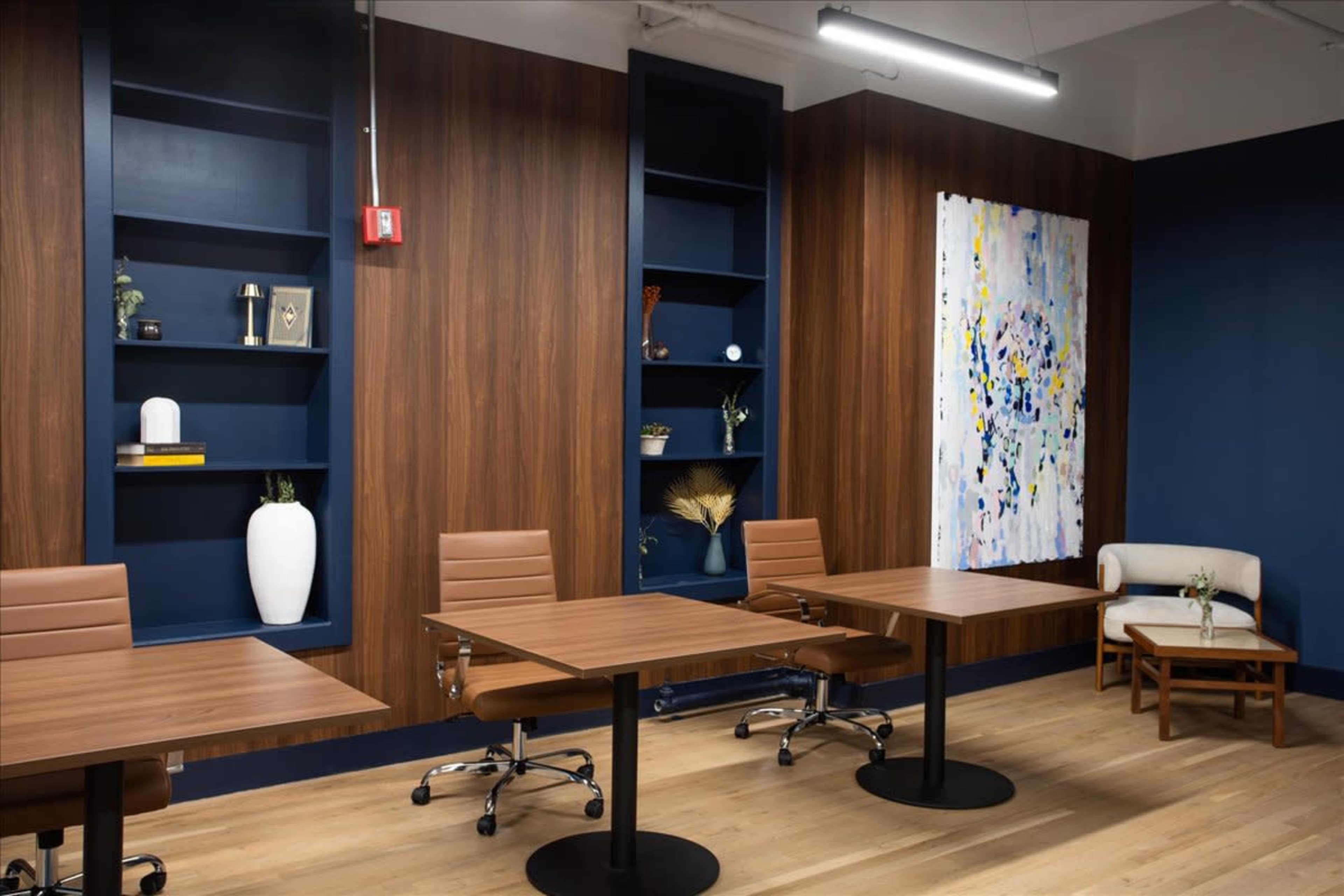 The image shows a modern conference room featuring wooden walls, two tables with rolling chairs, a minimalist couch, and decorative shelves.