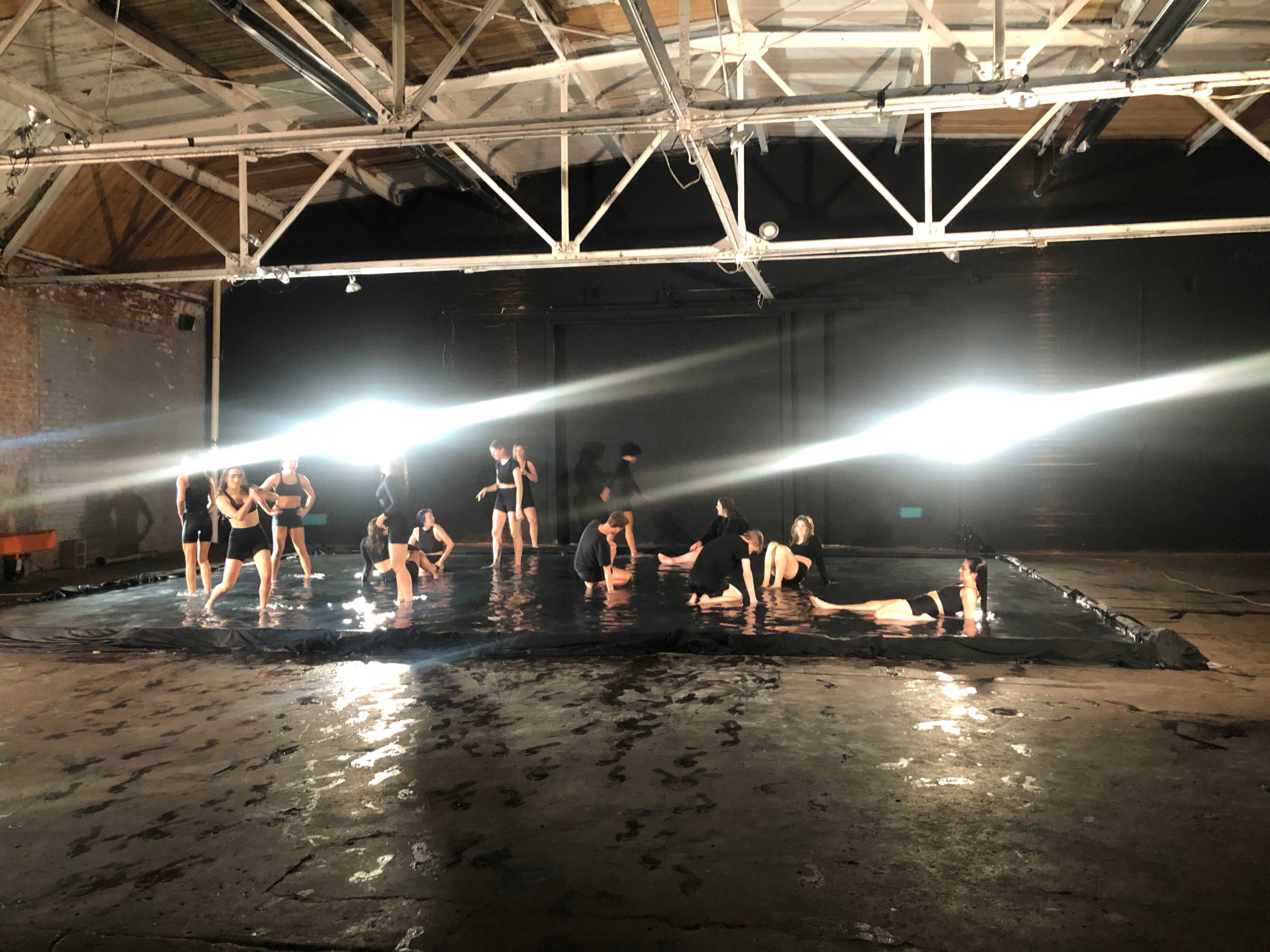 A group of individuals performs on a large black tarp in a dimly lit industrial space.