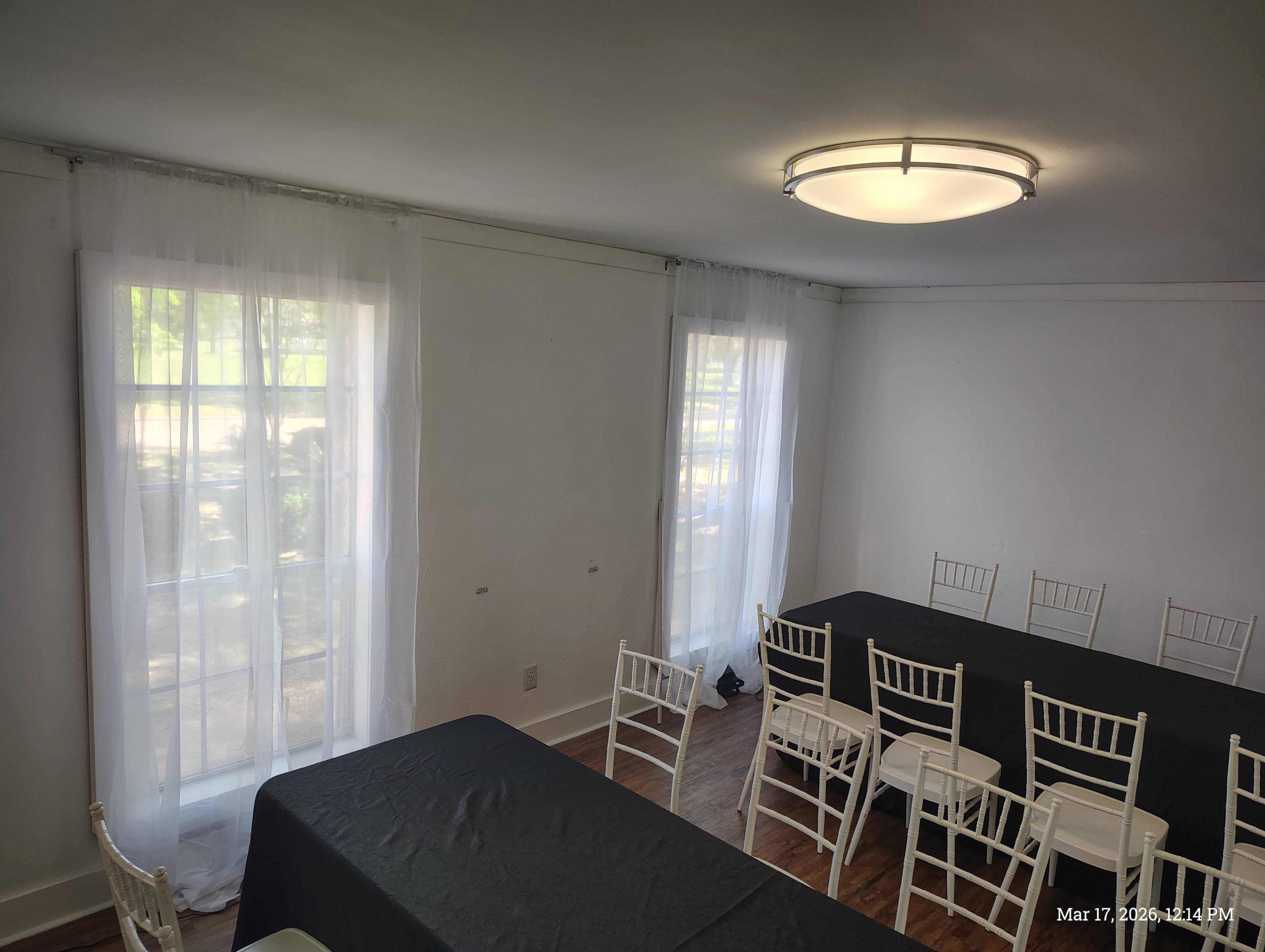 The image shows a bright, empty room with a large table set up with white chairs and sheer curtains framing two windows.