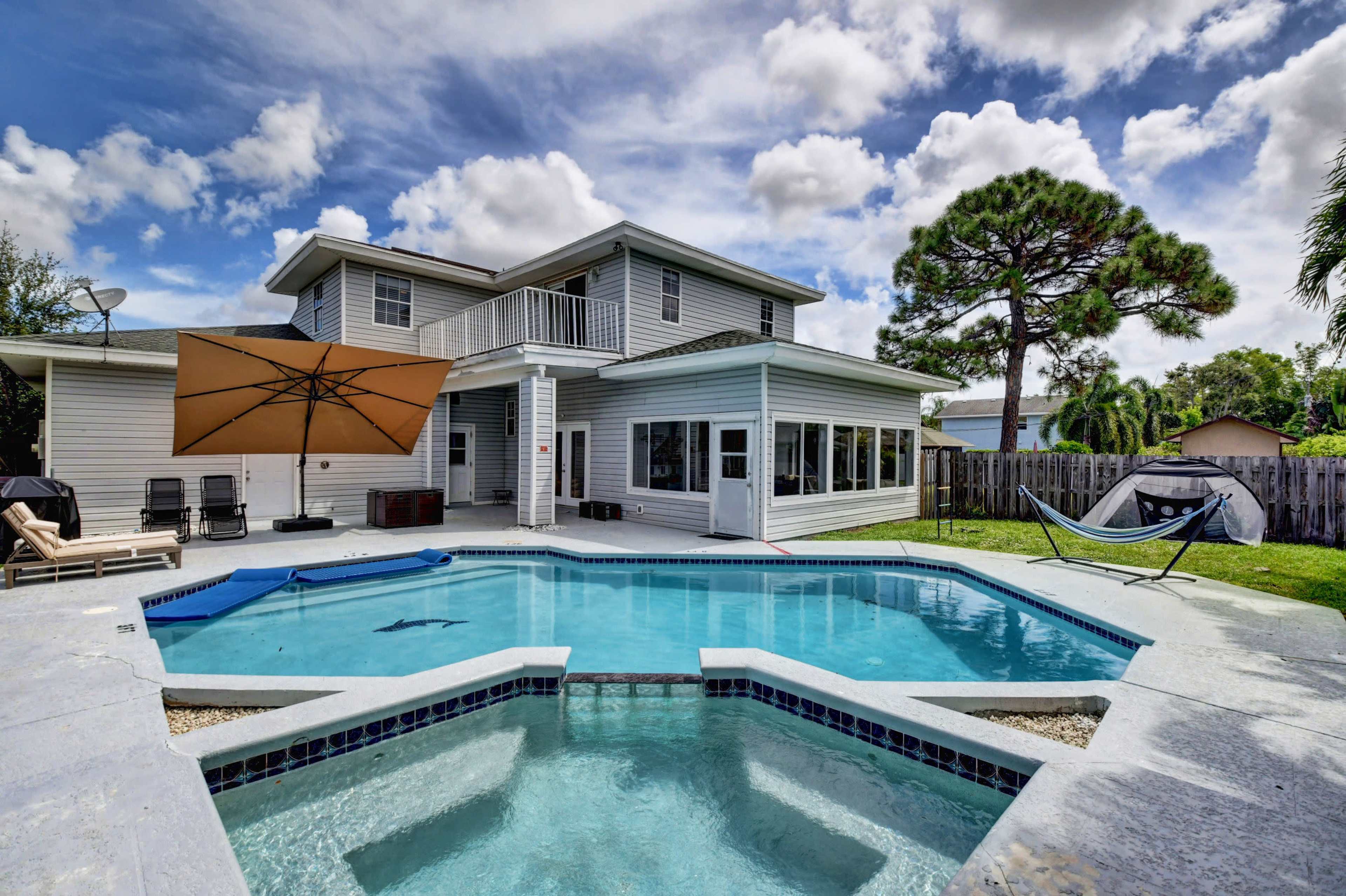 The image shows a two-story house with a swimming pool and a patio area featuring an umbrella and a hammock in the backyard.